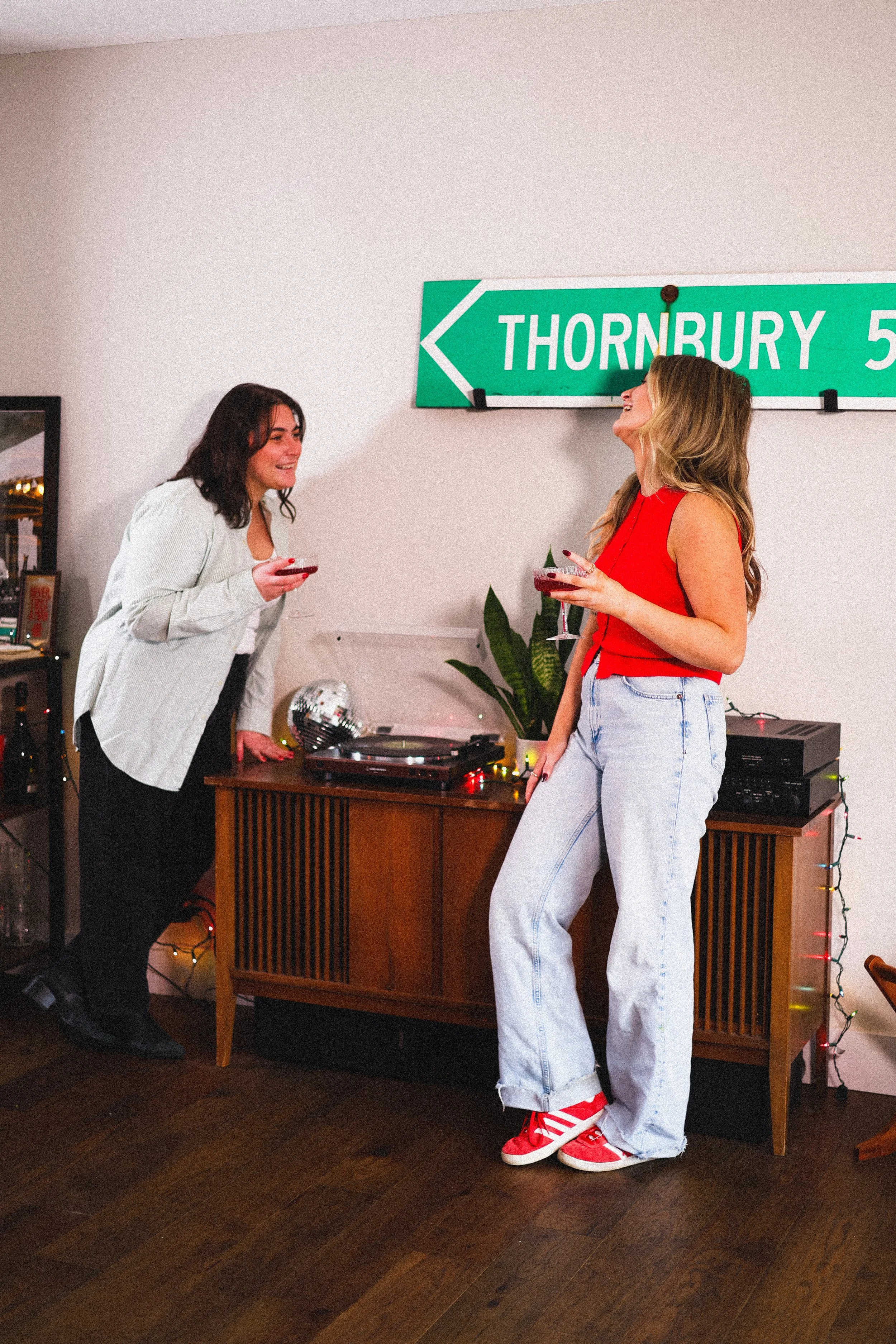 Two women converse and laugh at a social gathering, holding cocktails in a room decorated with string lights and a green street sign reading 'Thornbury 5' on the wall.