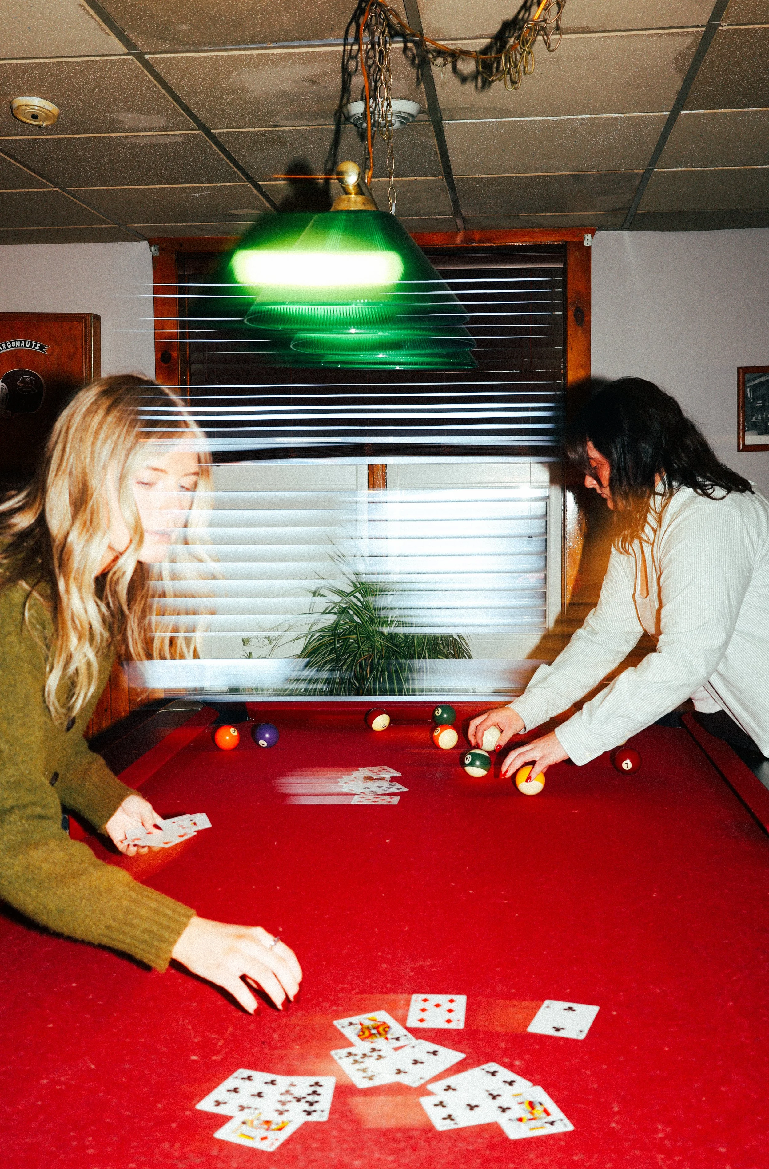 Two women playing pool, with cards on the table and pool balls positioned for a game, inside a room with a red pool table, a window, and a green hanging light.