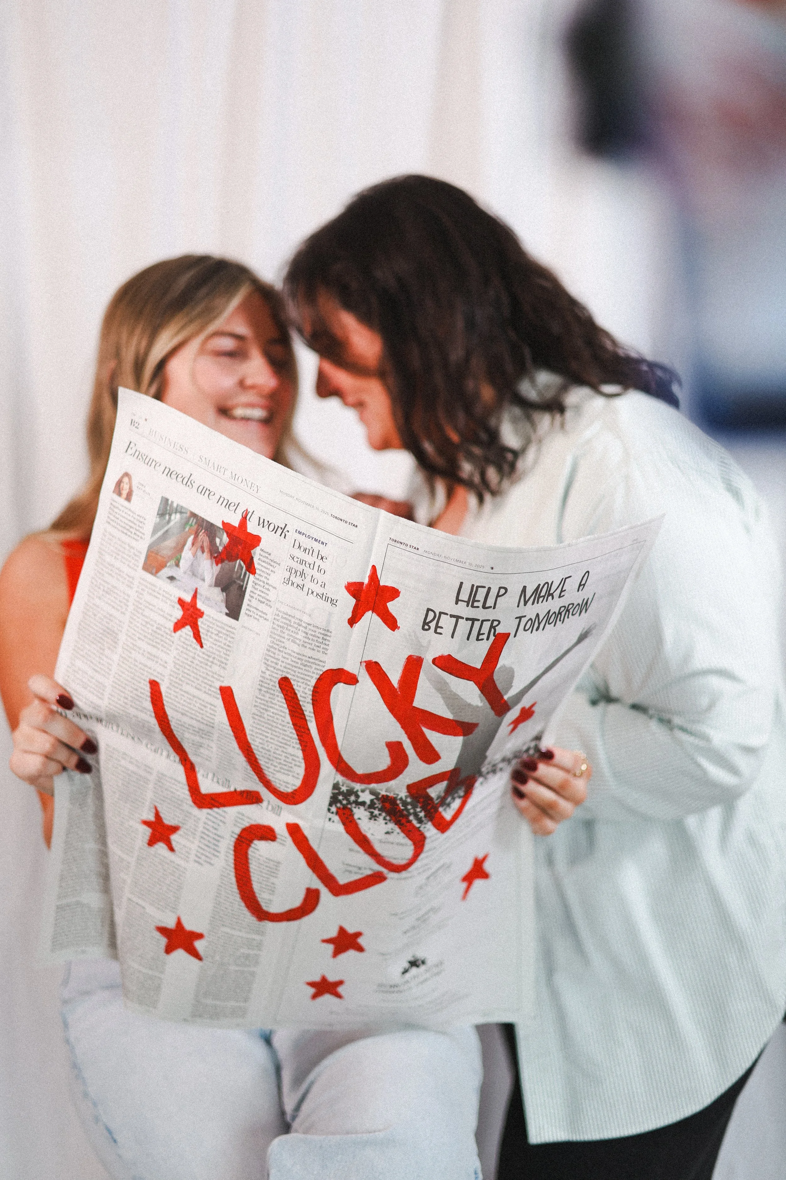 Two women are celebrating, one is sitting and holding a newspaper decorated with red stars and bold red text that reads 'LUCKY CLOSING'; the other woman is leaning toward her, smiling, with their foreheads touching.