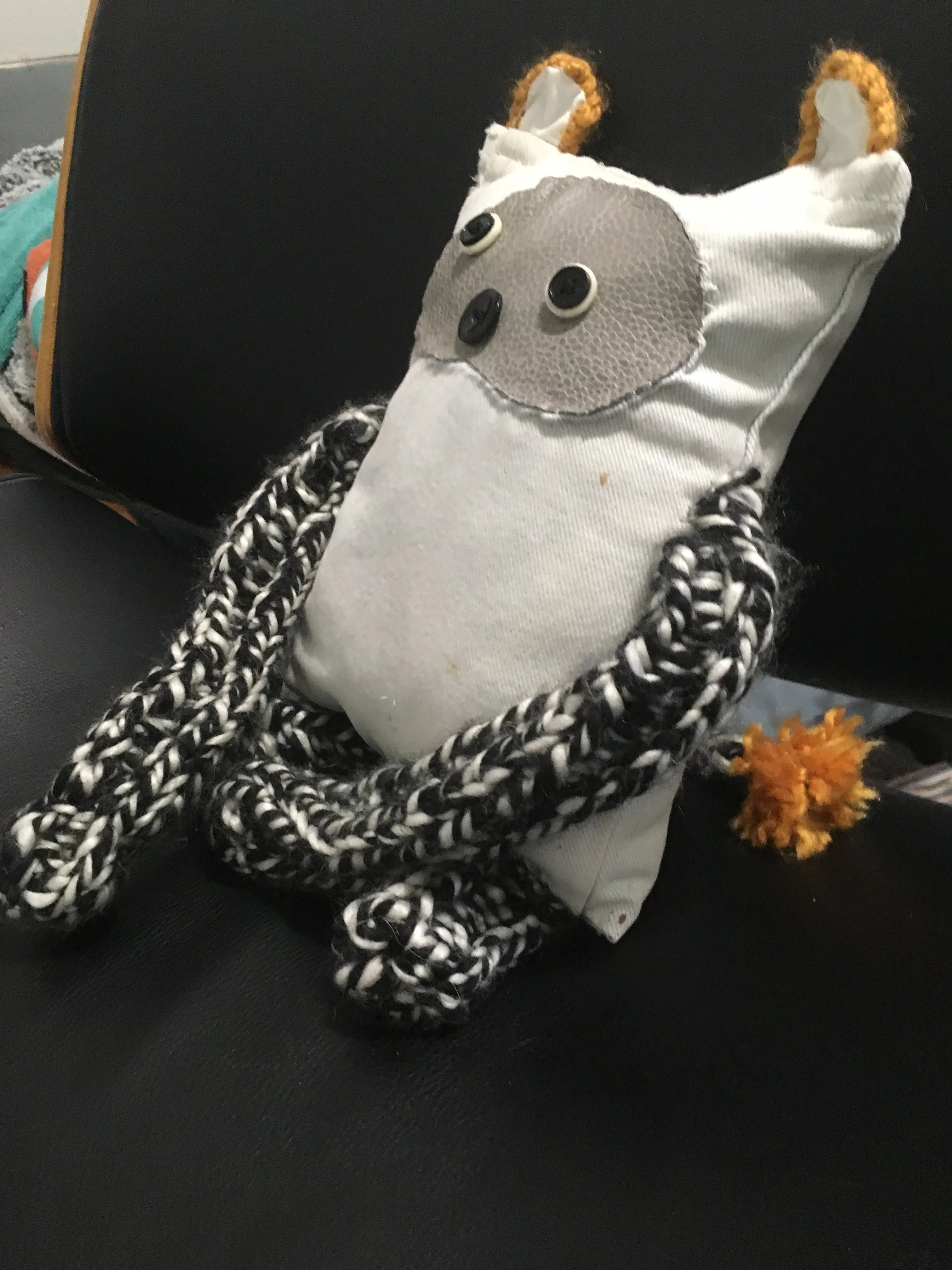 A plush toy resembling a bear with a white body, a beige circular face, button eyes, a button nose, black and white braided limbs, orange pom-pom tail, and ears made from white fabric with orange trim, resting on a black surface.
