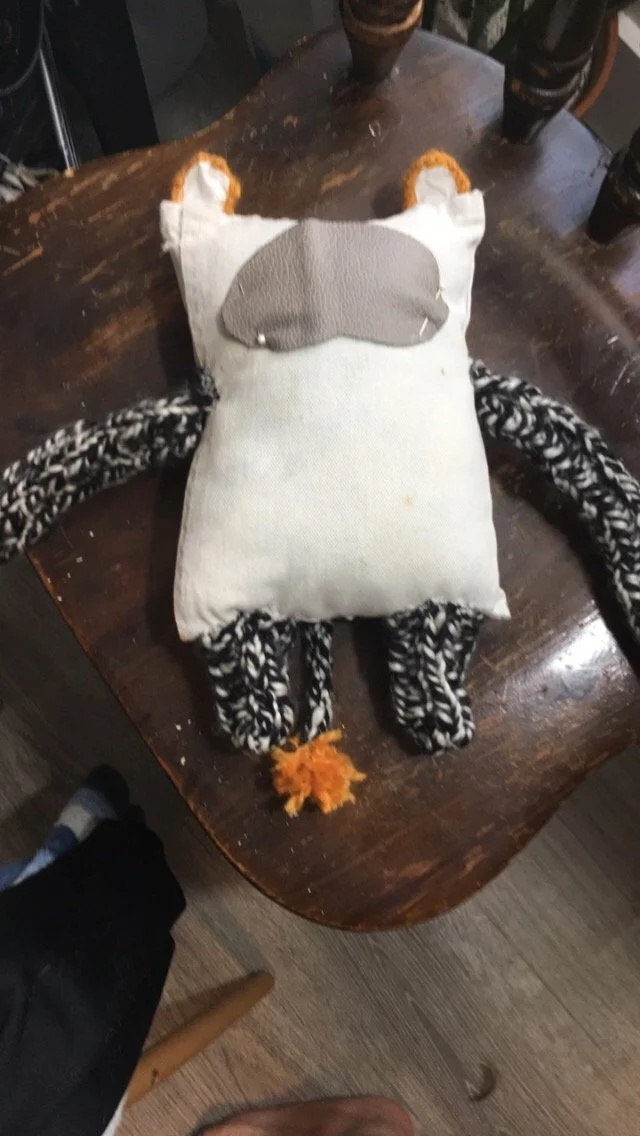 A stuffed owl toy with a white body, black and white patterned wings and legs, an orange pom-pom tail, and an owl face mask on the head, placed on a wooden table with cups around it.