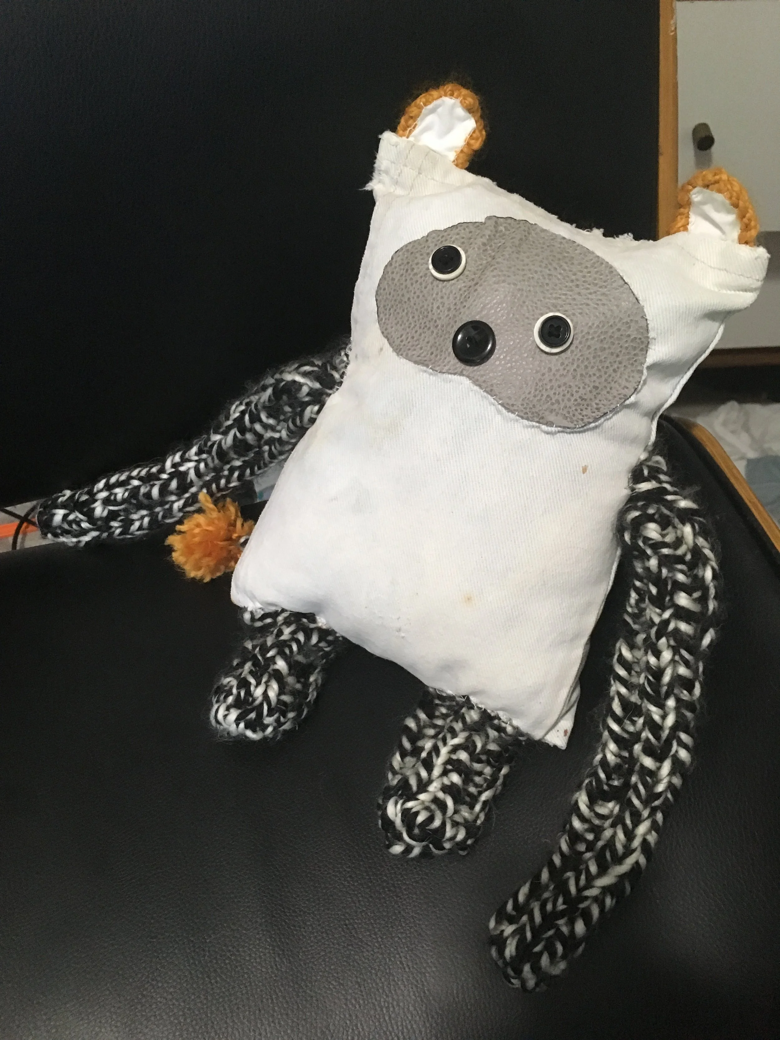 A stuffed toy resembling a cow with a fabric body, black and white braided arms and legs, and a gray face with button eyes.