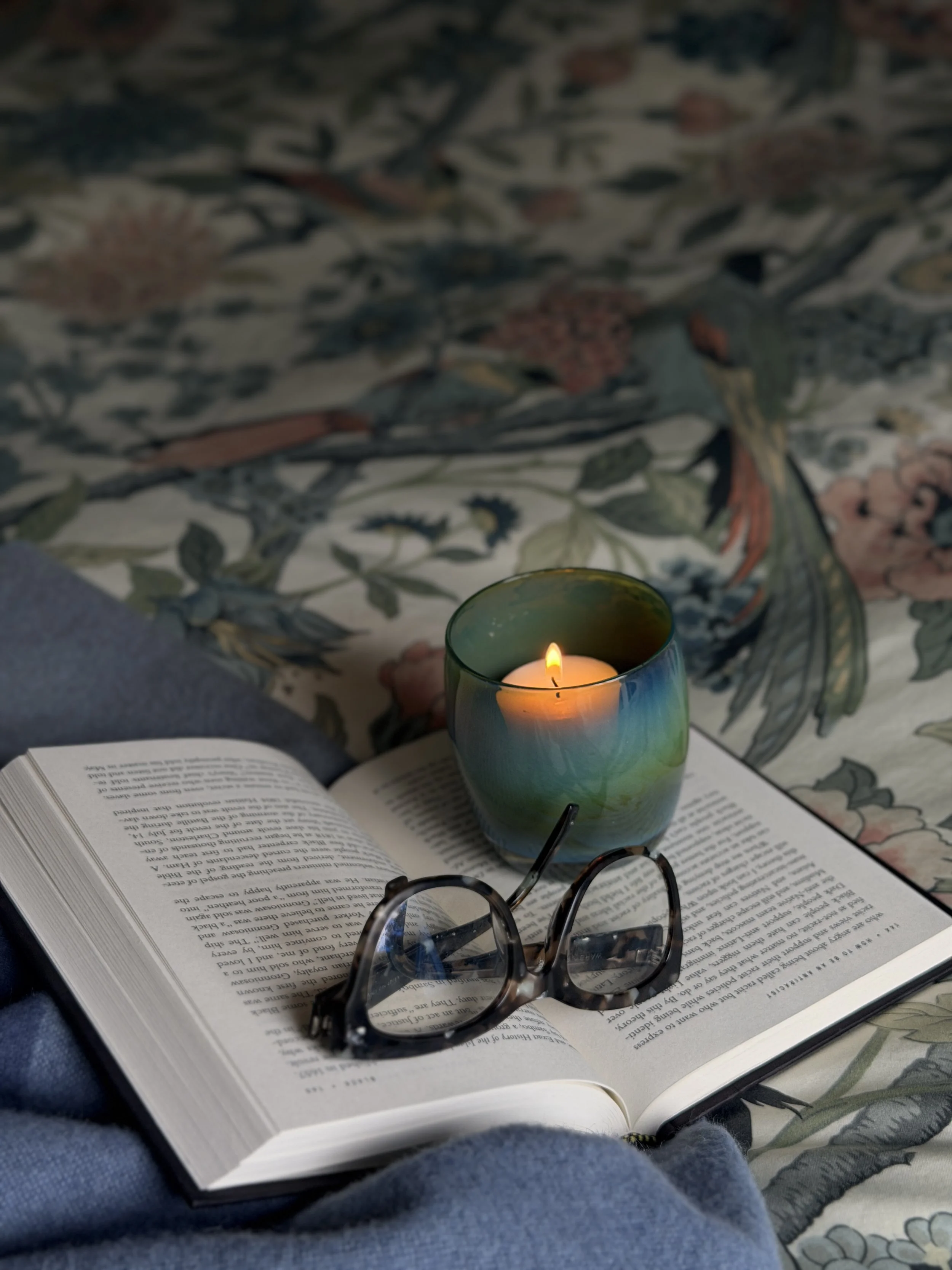 Open book, lit candle, and glasses on a bed.
