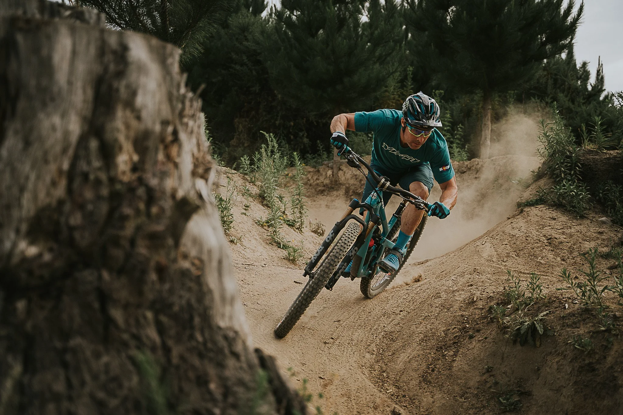 Jared Graves mountain biking in a turquoise shirt, shorts, and helmet riding down a dirt trail in a forest, kicking up dust.