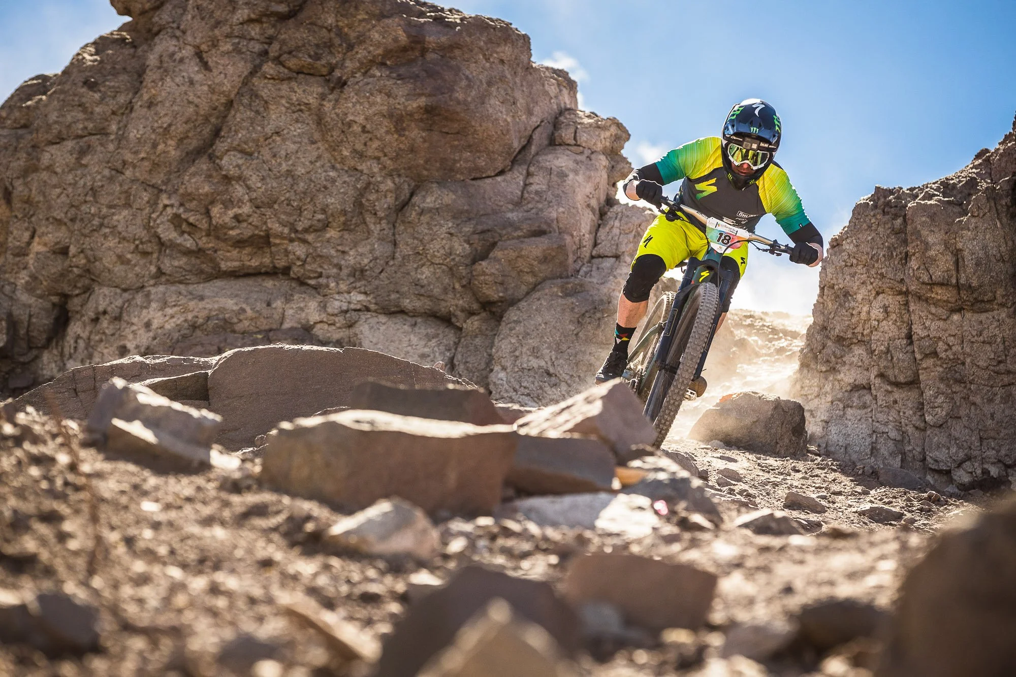A photo of Jared Graves, mountain biking in a black, yellow, and green race wear with a full-face helmet and goggles riding down a rocky, dusty trail between large rocks and boulders under a clear blue sky.