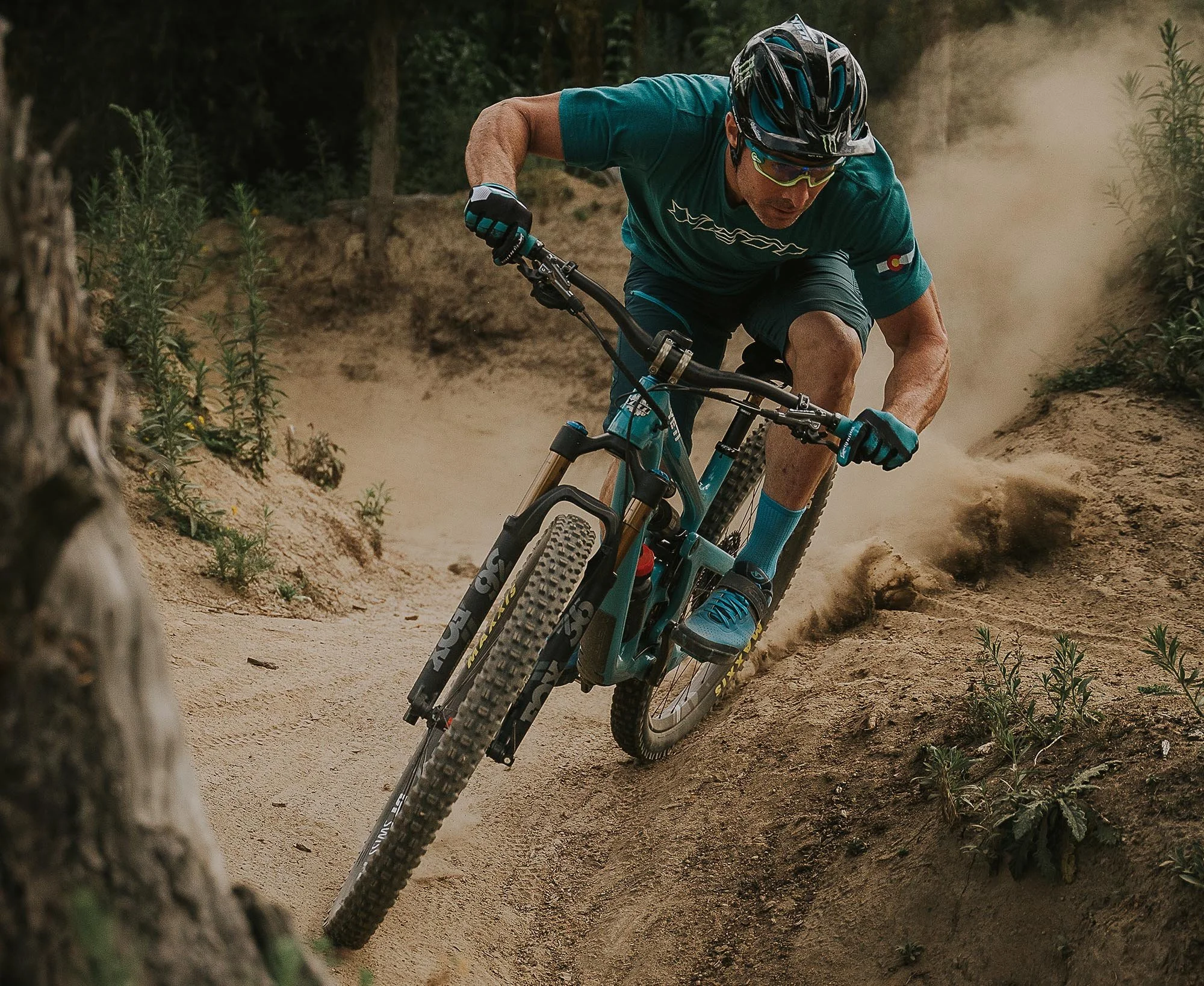 A photo of Jared Graves Mountain biking on a dirt trail in a forested area, wearing a helmet, sunglasses, gloves, and turquoise sportswear, a Yeti Cycles bike, leaning into a turn with dust kicking up behind him.