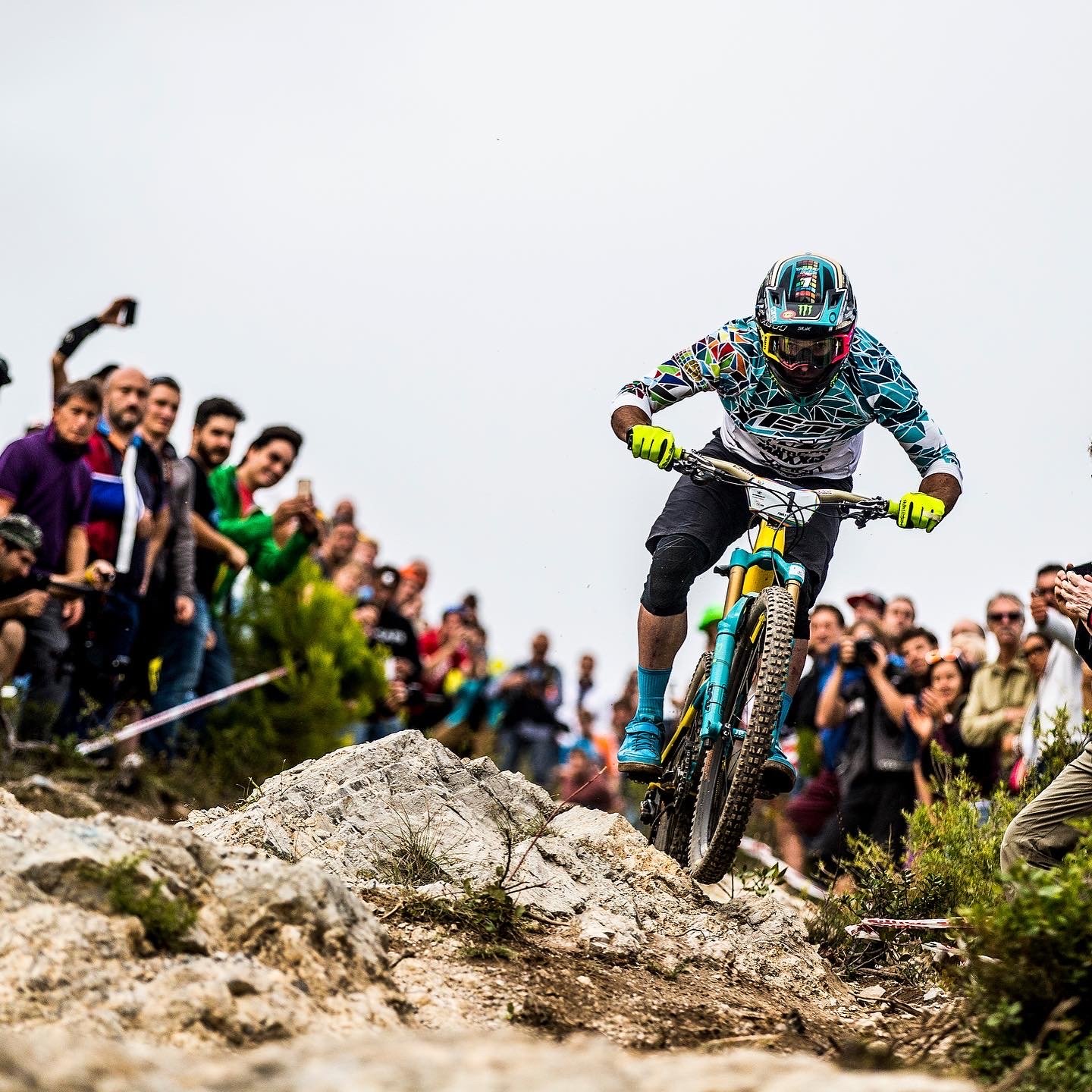A mountain biker wearing a helmet, goggles, and colorful gear rides downhill on rocky terrain as a crowd of spectators watches and takes photos in the background.