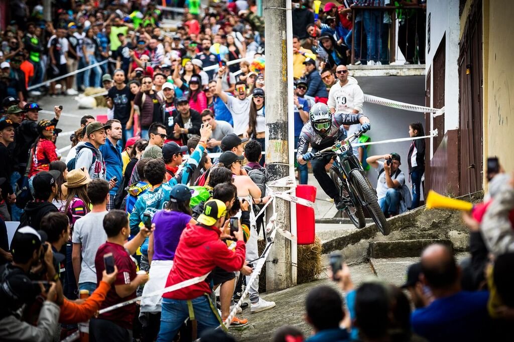 A photo of Jared Graves Mountain biking in a full-face helmet, goggles and gloves, riding down an urban street with a crowd of spectators cheering and taking photos. The scene shows a steep concrete staircase and many people gathered along the sides.