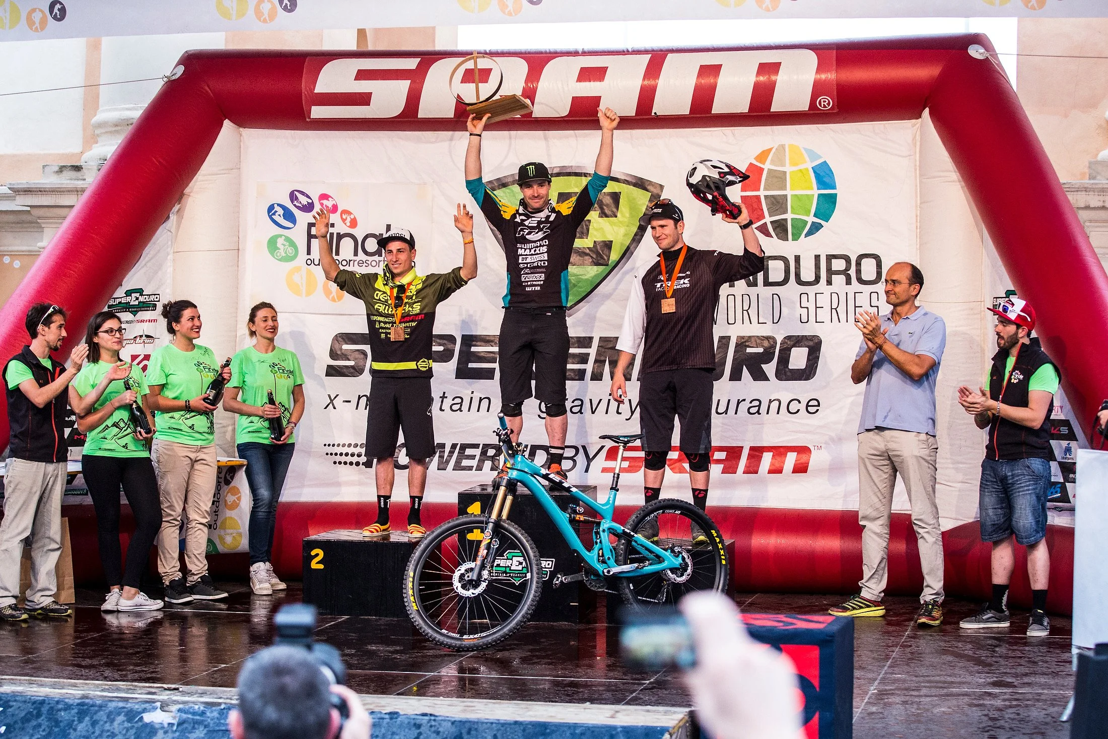 Enduro World Cup Mountain biking podium with three male winners, the first-place rider is Jared Graves, they are surrounded by officials and spectators, at an Enduro mountain bike race event.