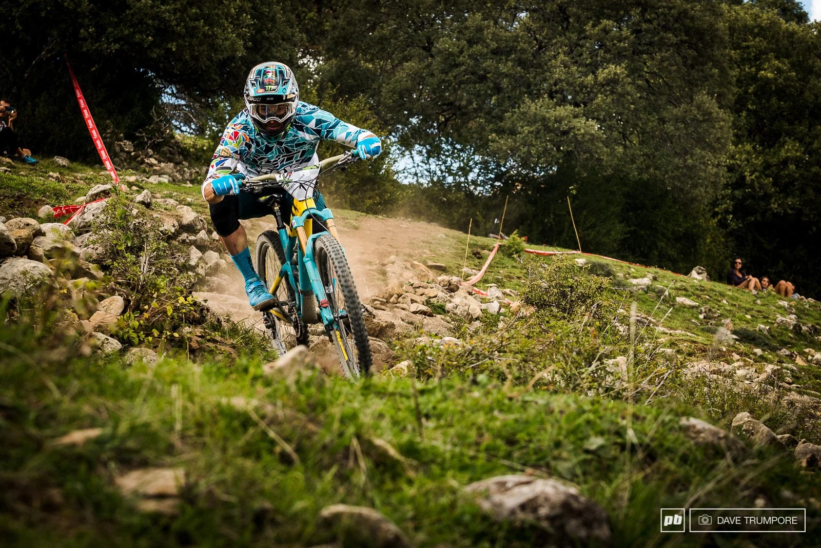 A mountain biker riding downhill on a rocky trail, wearing a helmet and colorful gear, with spectators sitting on the hillside in the background.