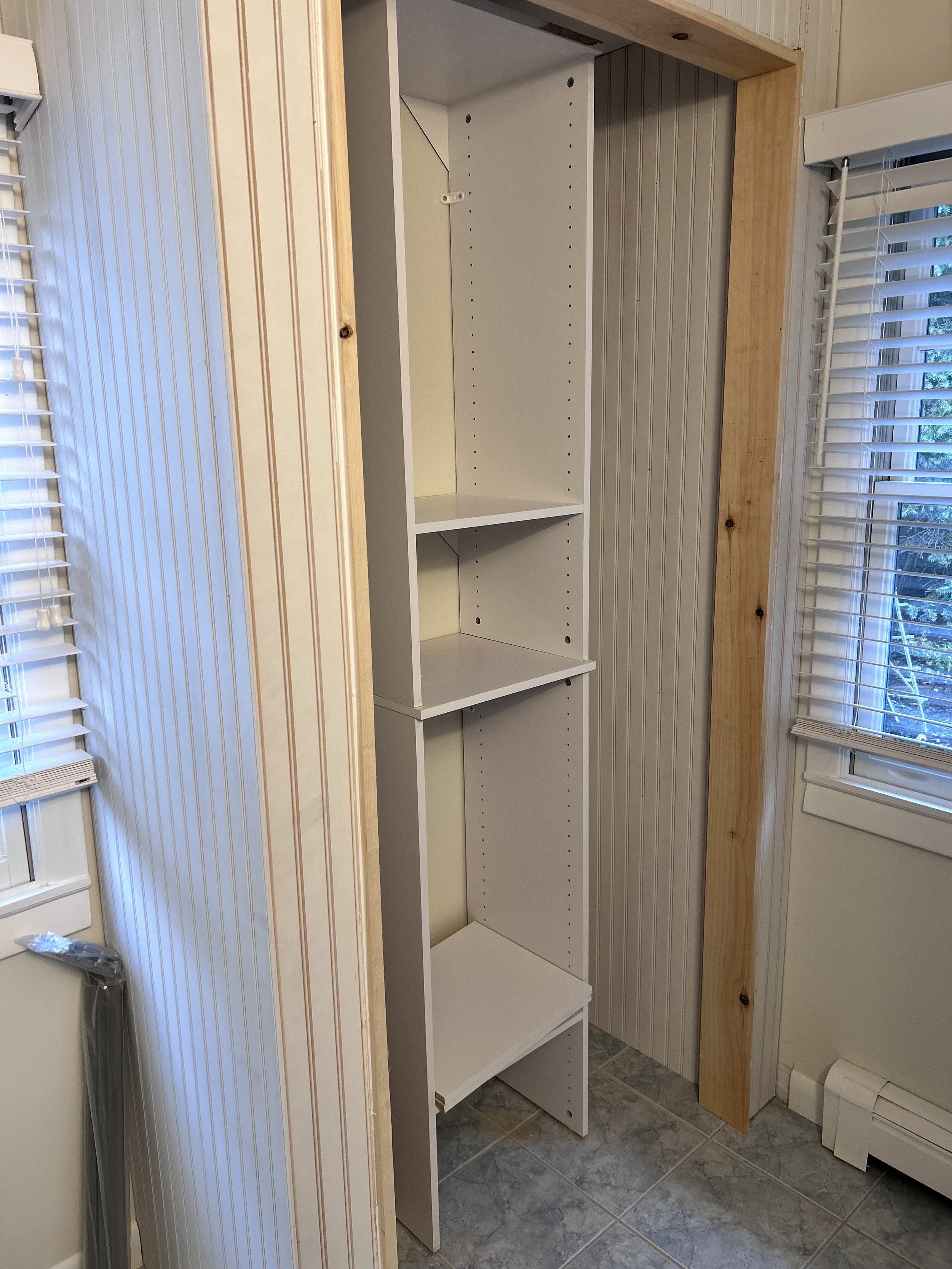 An unfinished tall, white shelving unit installed between two walls, with four shelves inside. The room has wood-paneled walls and a tile floor, with windows and blinds on either side of the shelving.