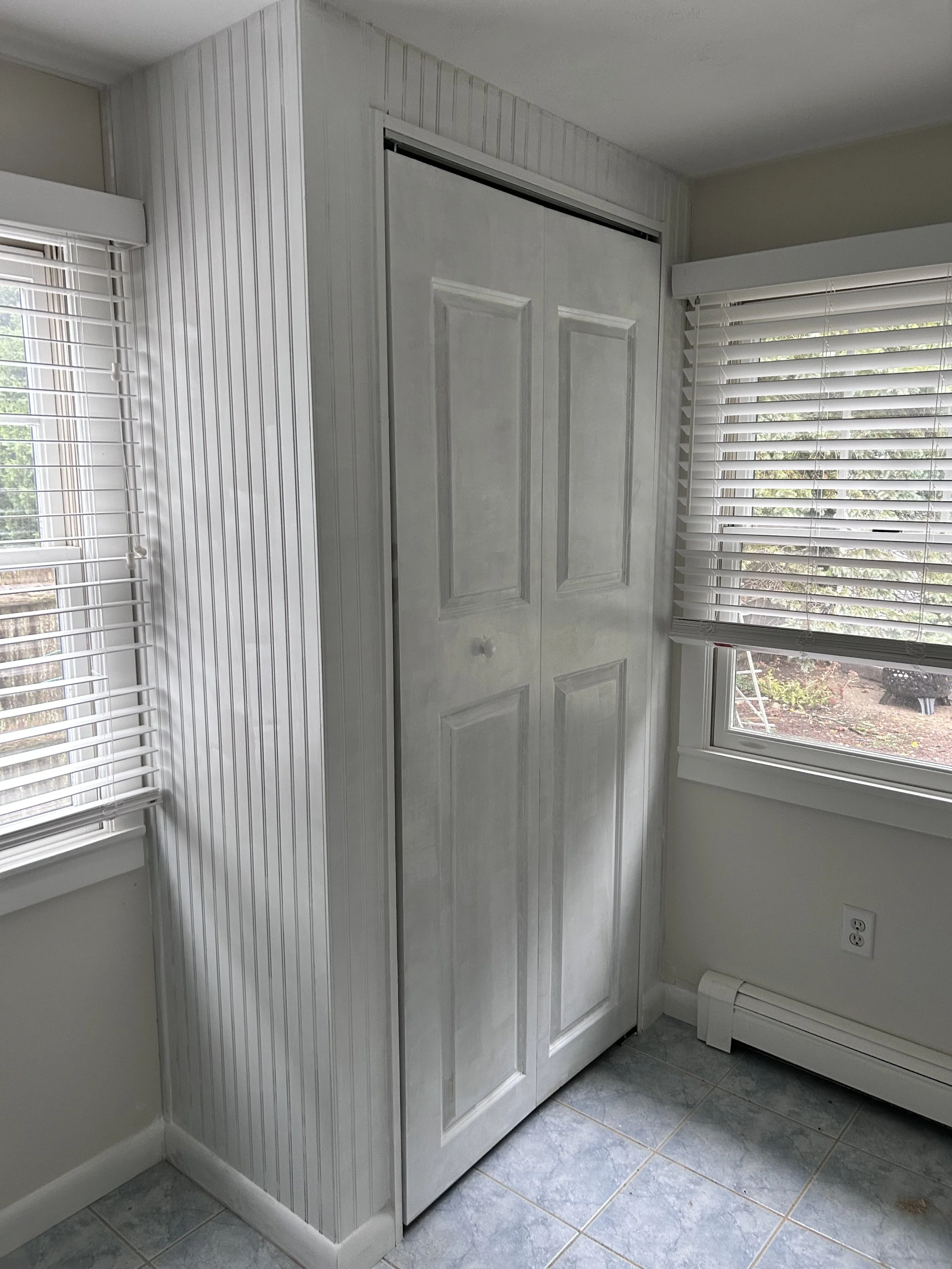 White closet with sliding doors in a room with two windows and blinds, a white wall, and tiled floor.