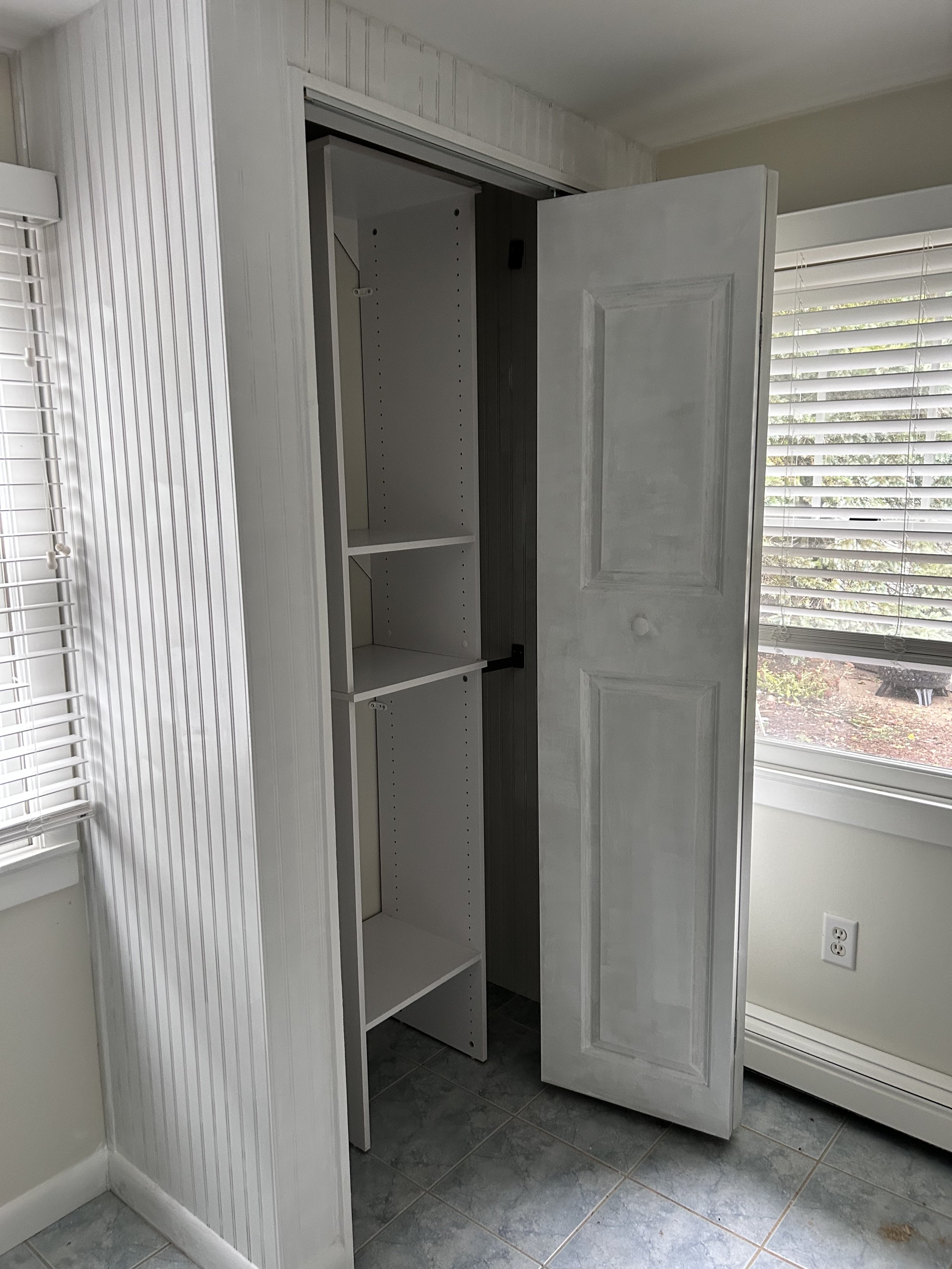 Empty closet with adjustable shelves, partially open door, in a room with a window and blinds, tiled floor, and a wall outlet nearby.