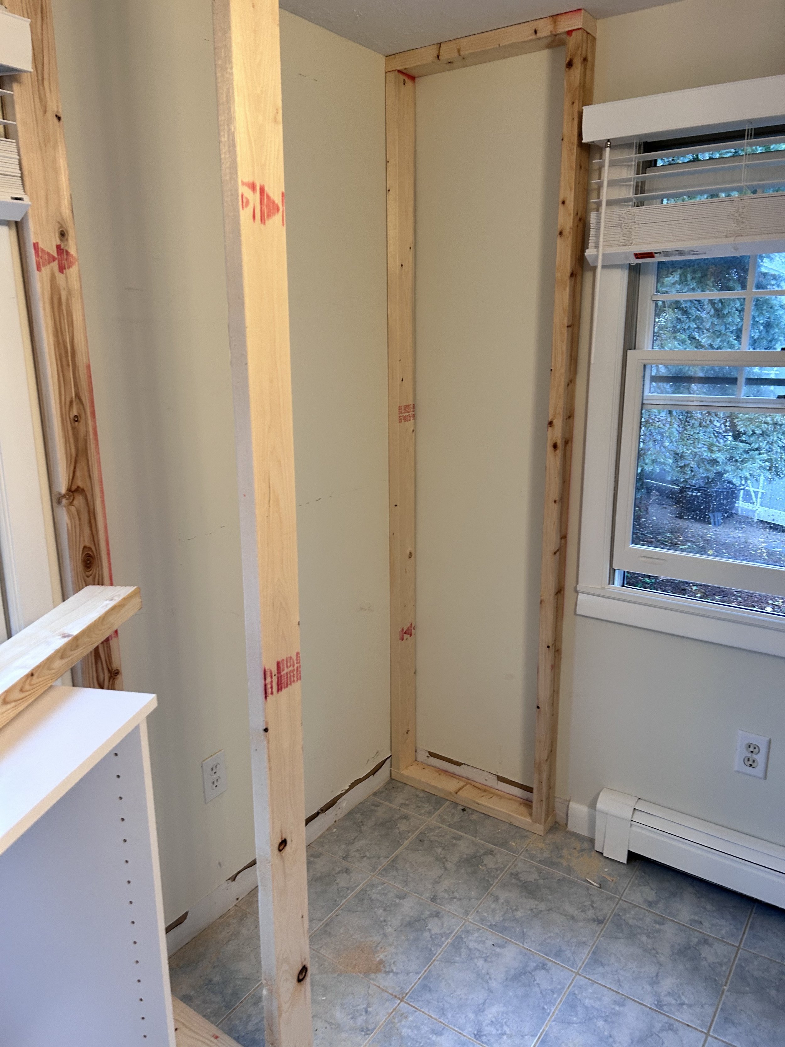 Interior of a room with wooden closet framing, a window with blinds, a white baseboard heater, and partially installed shelving or wall.