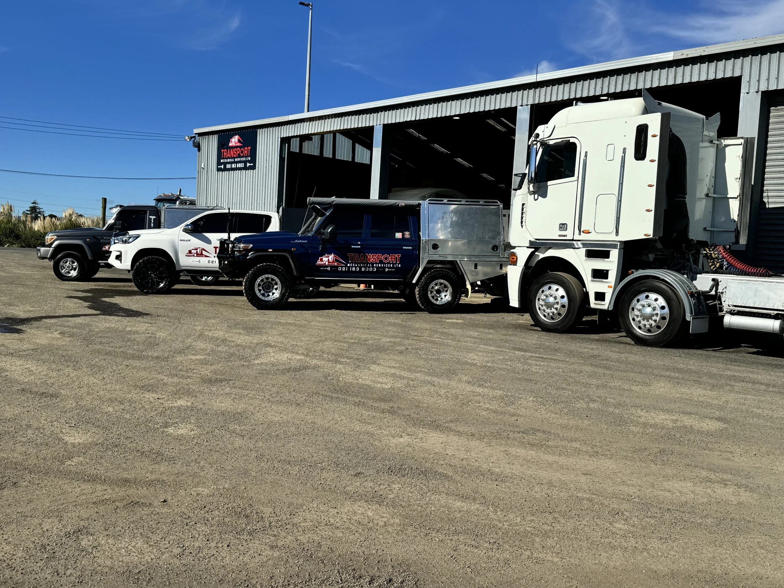 A fleet of vehicles parked outside workshop, including a large white semi-truck, 3 utes with sign writiing.