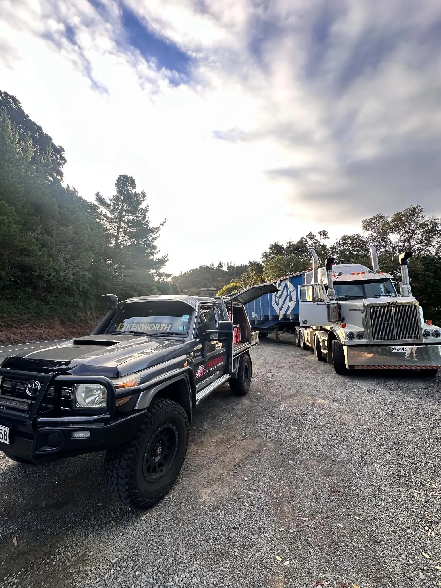 A dirt road on a hillside with sparse trees and bushes, featuring an orange excavator and a  vehicle parked near each other.
