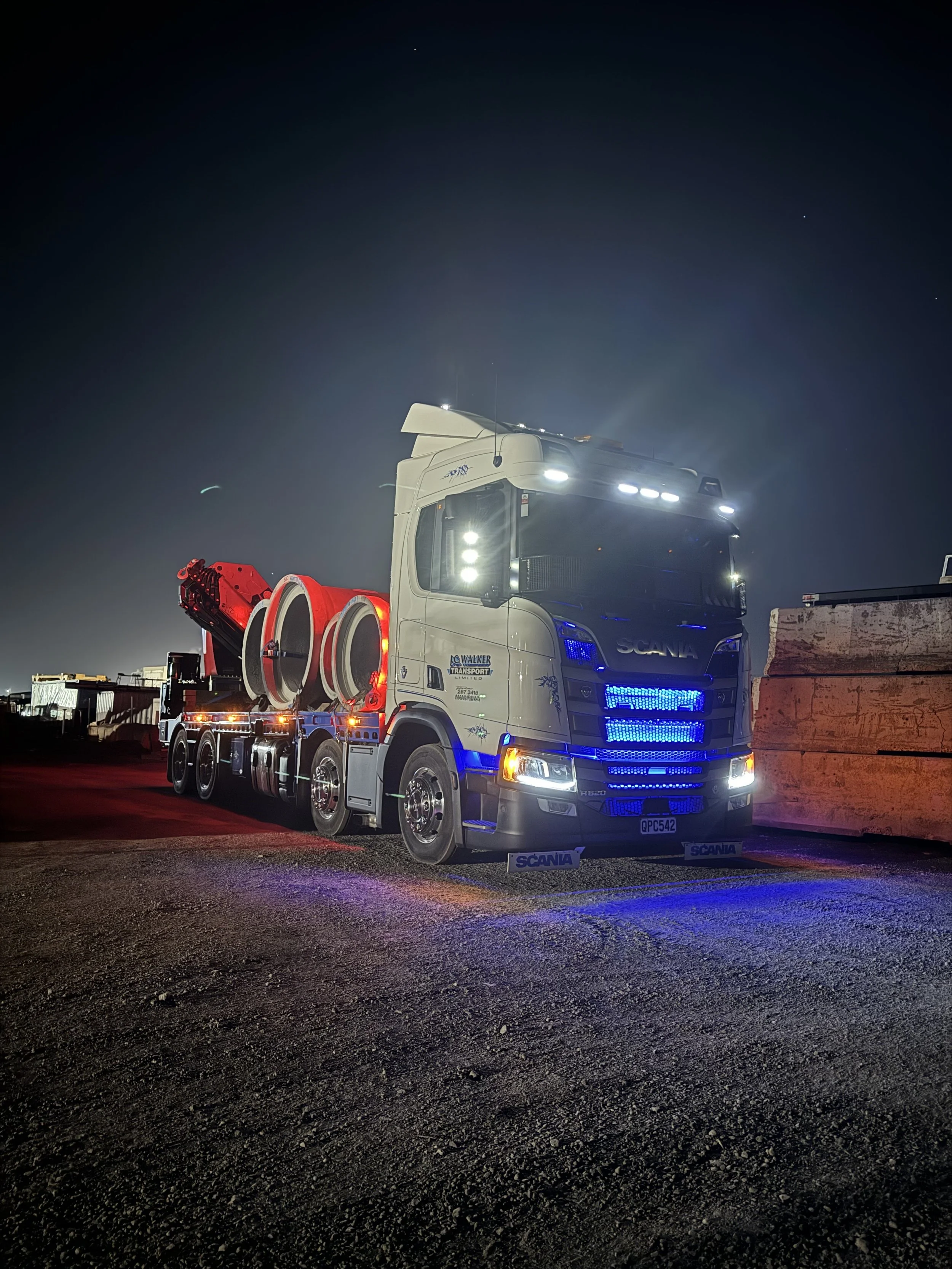 A large white semi-truck with bright lights parked outdoors at night, with some large wooden pallets visible on the right side.