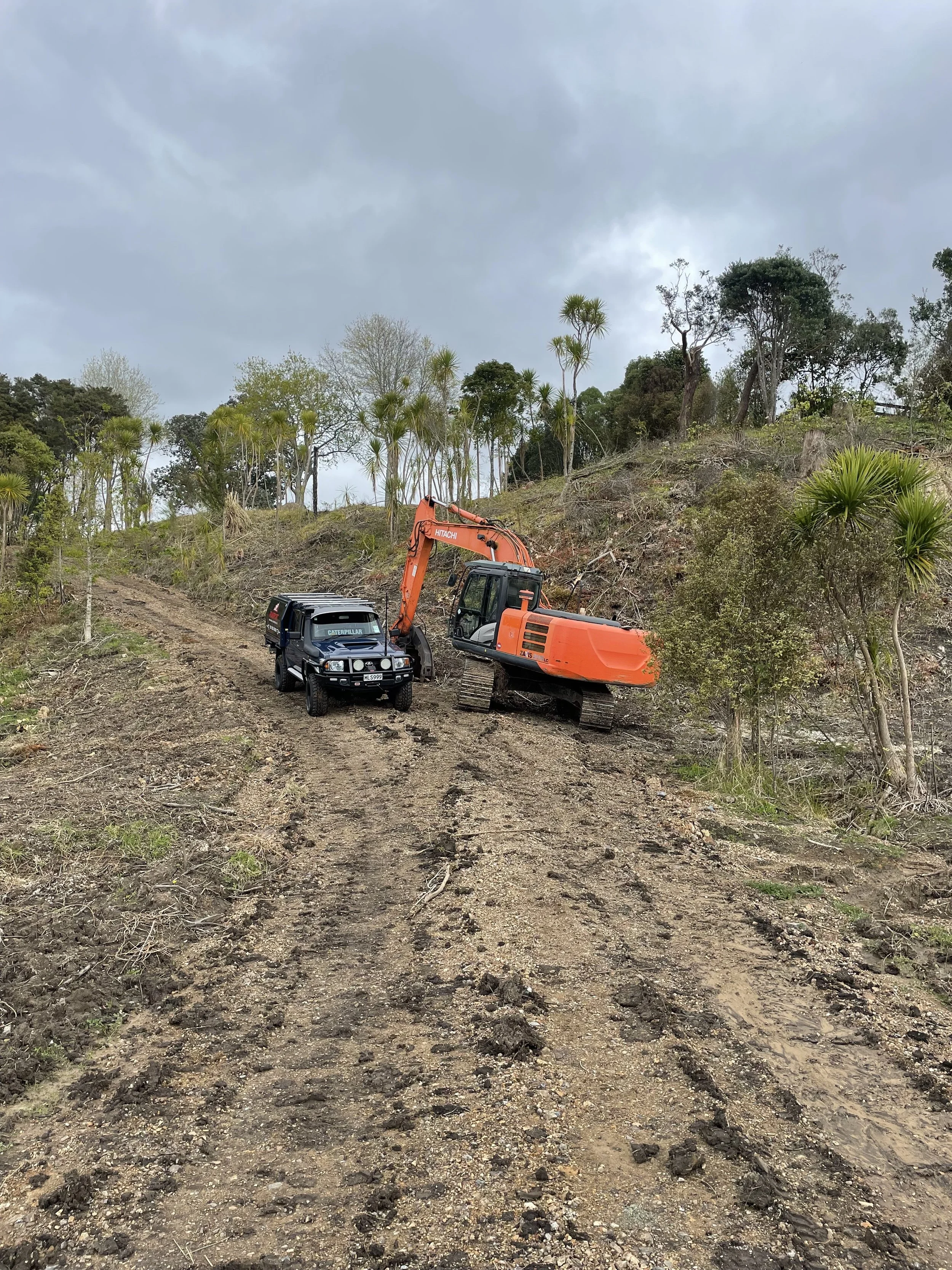 A dirt road on a hillside with sparse trees and bushes, featuring an orange excavator and a  vehicle parked near each other.