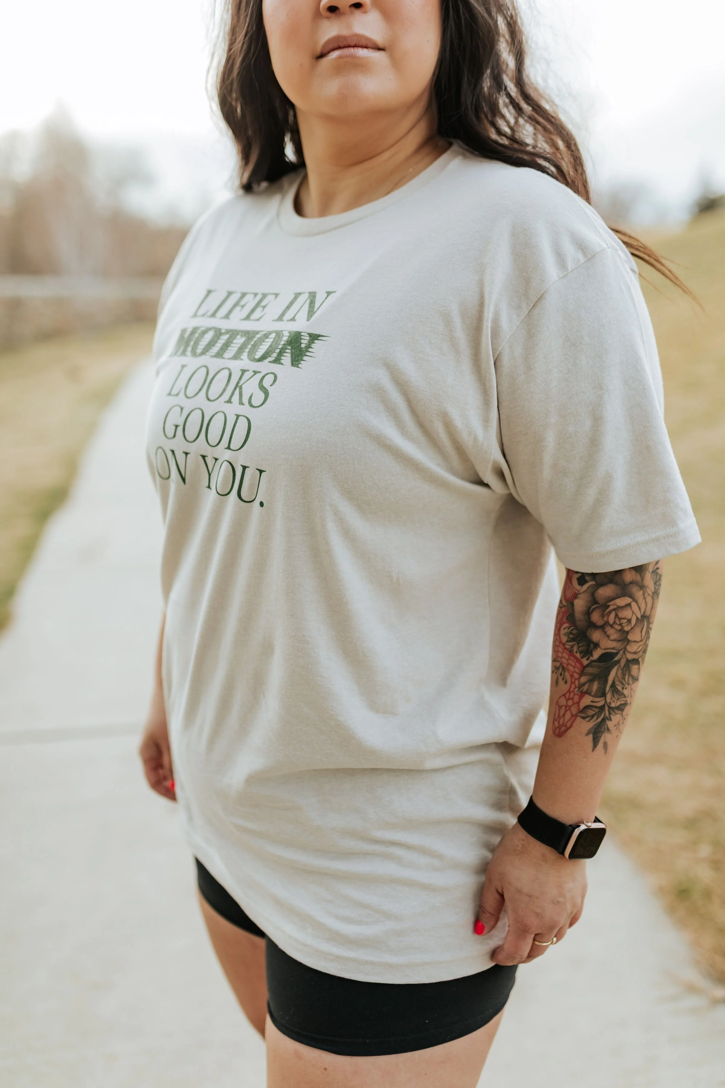 A woman outdoors wearing a beige T-shirt with the quote 'LIFE IN MOTION LOOKS GOOD ON YOU' in green letters, black shorts, a black smartwatch, and a floral tattoo on her left arm.