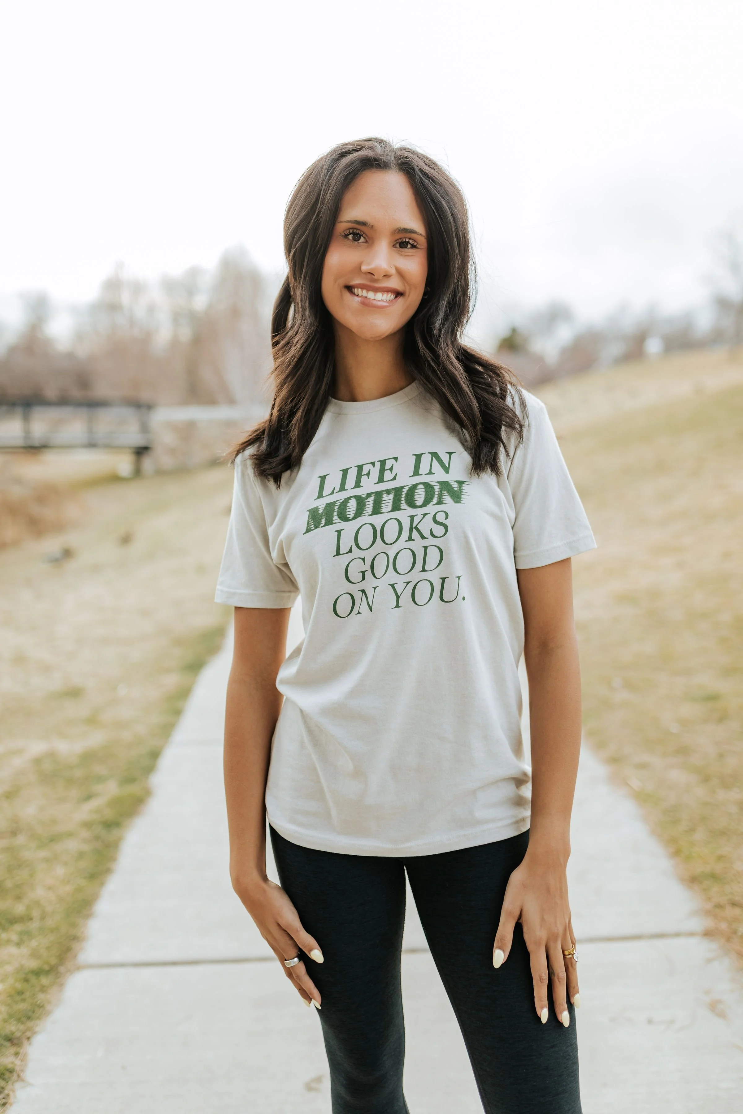 A young woman with dark hair wearing a white t-shirt with green text that says Life in Motion looks good on you, and wearing black leggings, standing outdoors on a sidewalk with a grassy area and trees in the background, smiling at the camera.