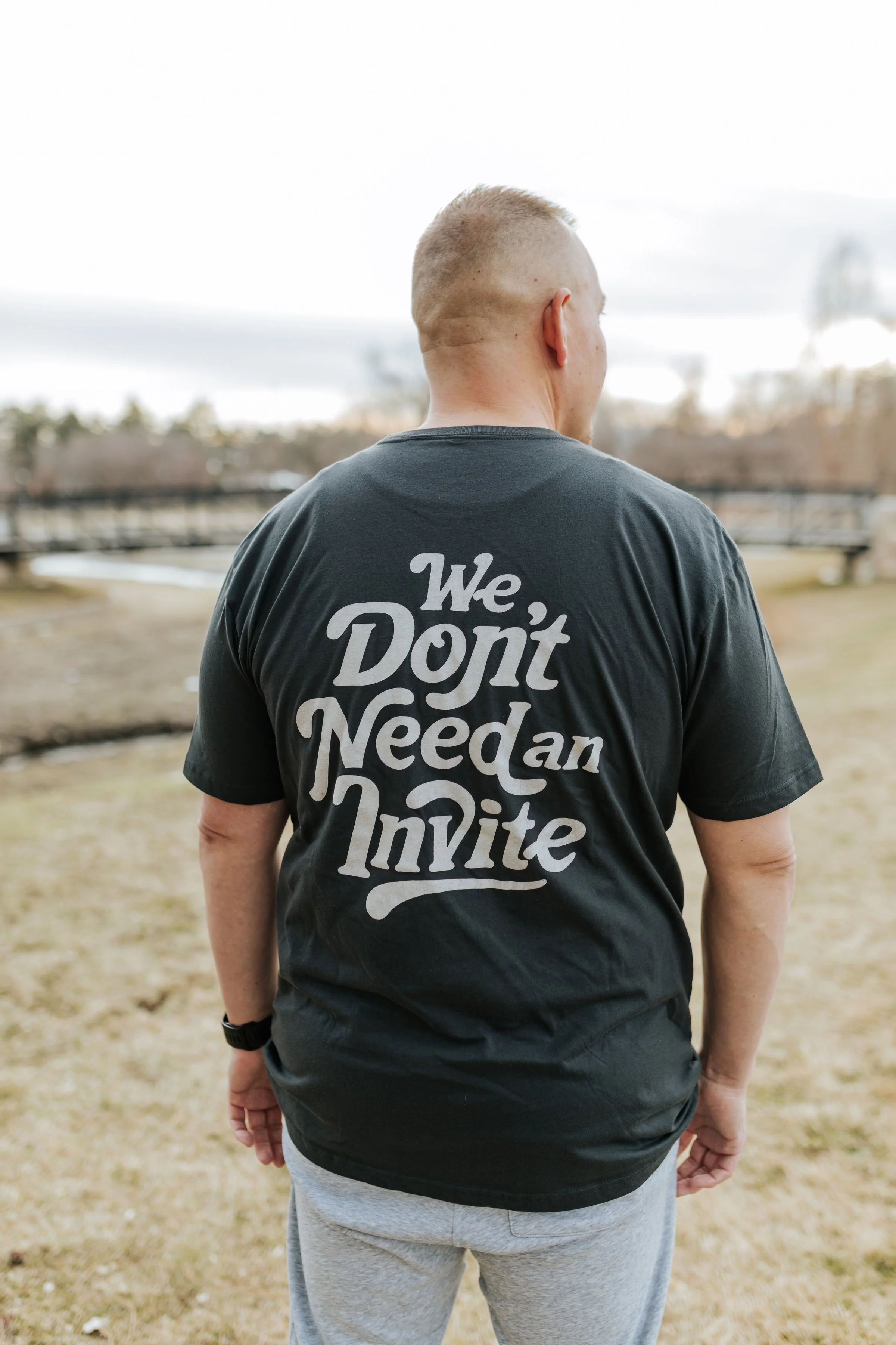 Back of a person wearing a black T-shirt with a message that says "We Don't Need an Invite" printed on it, outdoors with a blurred background including a bridge, trees, and water.