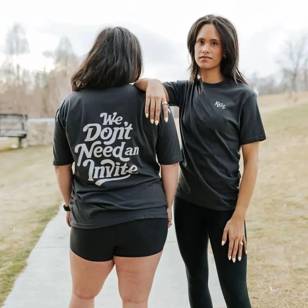Two women standing outdoors on a cloudy day, one with her back to the camera wearing a black t-shirt with the message 'We Don't Need an Invite', and the other facing the camera with a serious expression wearing a black t-shirt with "KM"  on the front