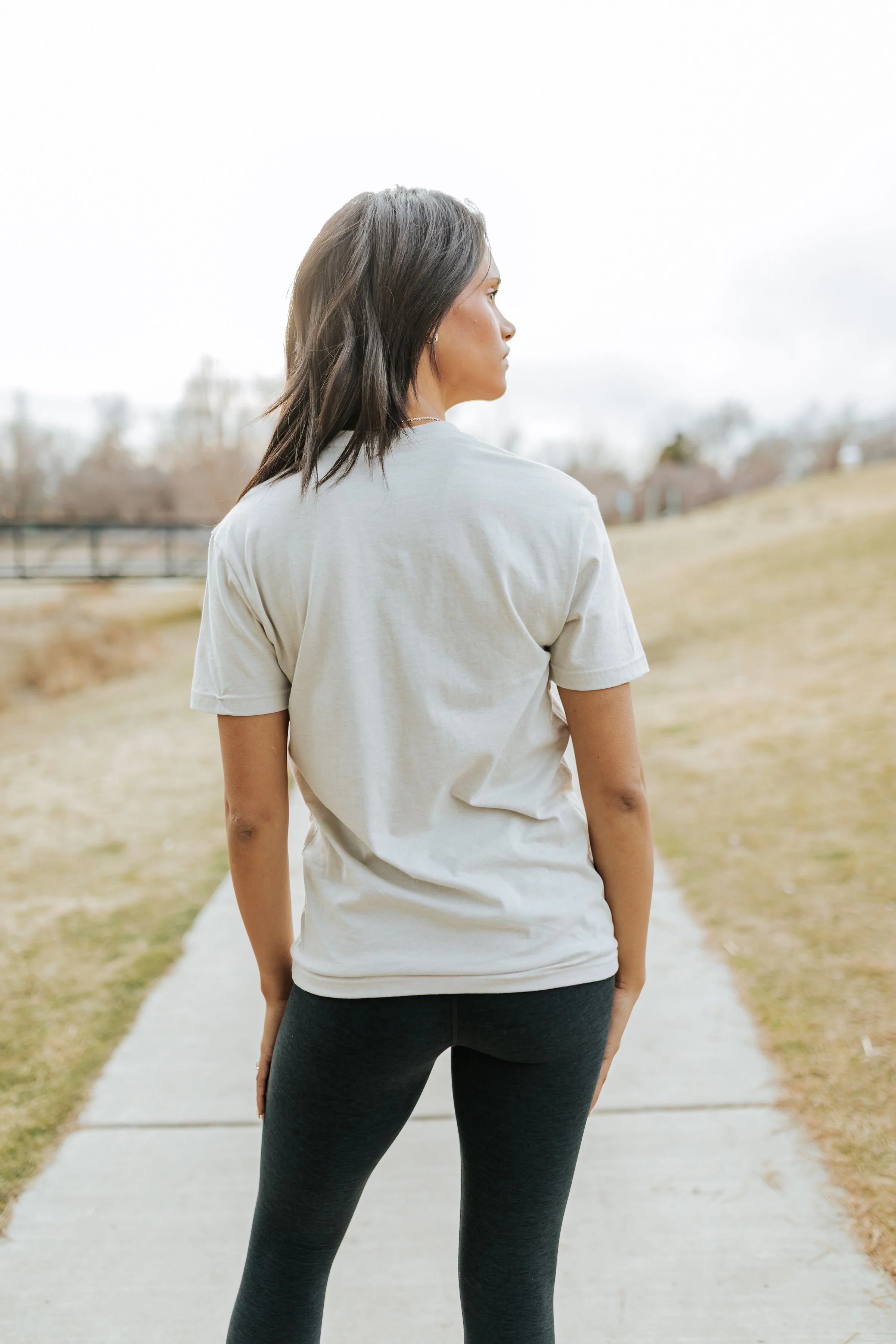 A woman standing on a sidewalk outdoors, facing sideways with her head turned slightly to the left, wearing a white t-shirt and dark leggings.