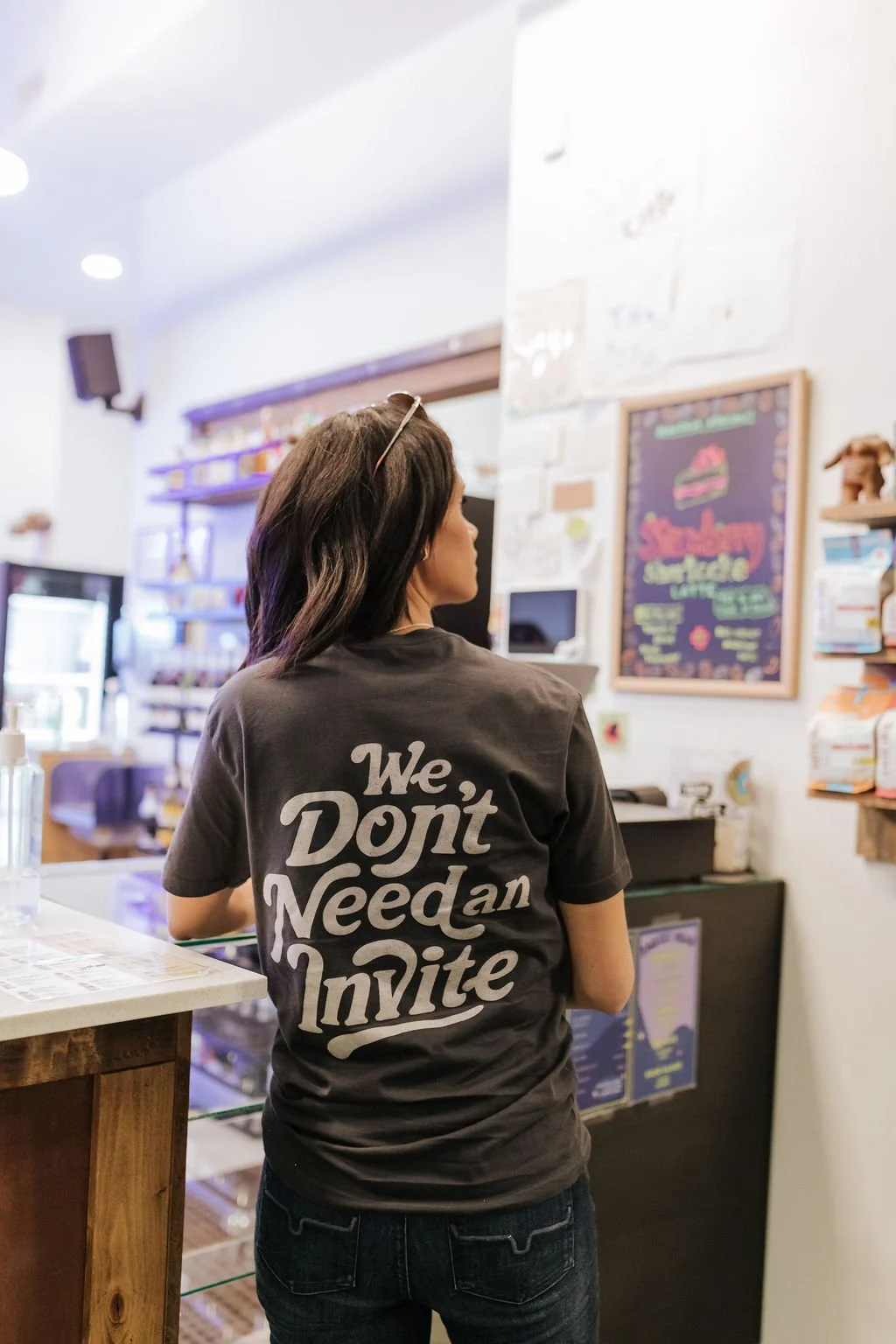 A woman with dark hair wearing a black T-shirt with white text that says, 'We Don't Need an Invite,' standing in a cafe.
