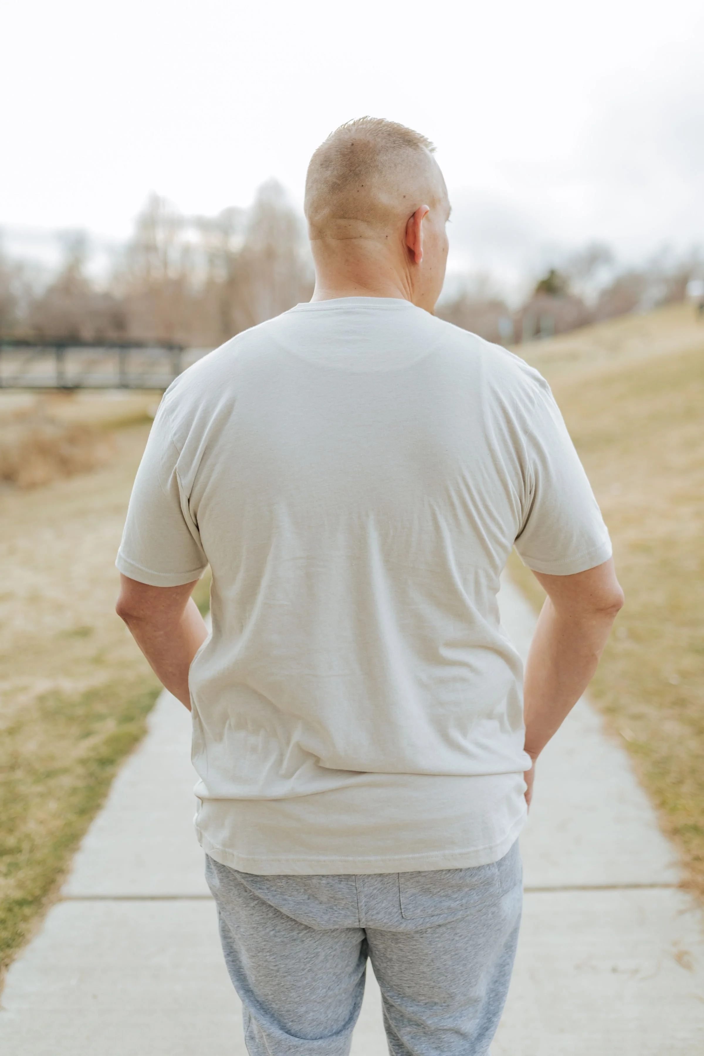Back view of a man with short blond hair, wearing a light gray T-shirt and gray sweatpants, standing on a sidewalk in an outdoor park area.