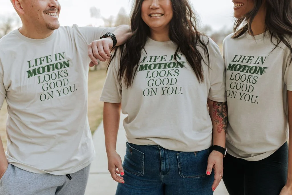 Three people wearing matching beige T-shirts that say "Life in Motion looks good on you" with green text, standing outdoors and smiling.