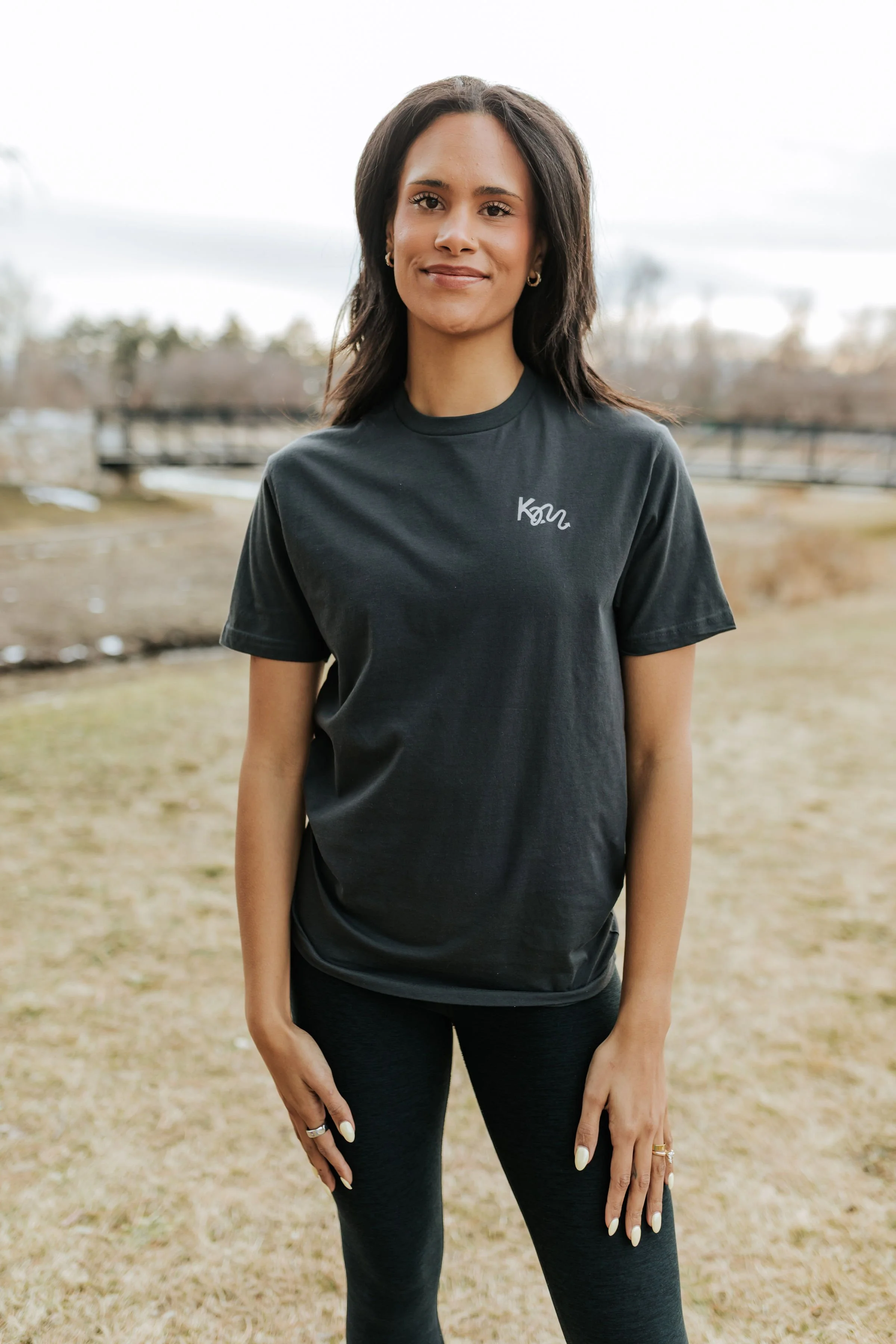 A young woman standing outdoors in a field, wearing a black T-shirt with the word 'KM' on it and black leggings, smiling at the camera.