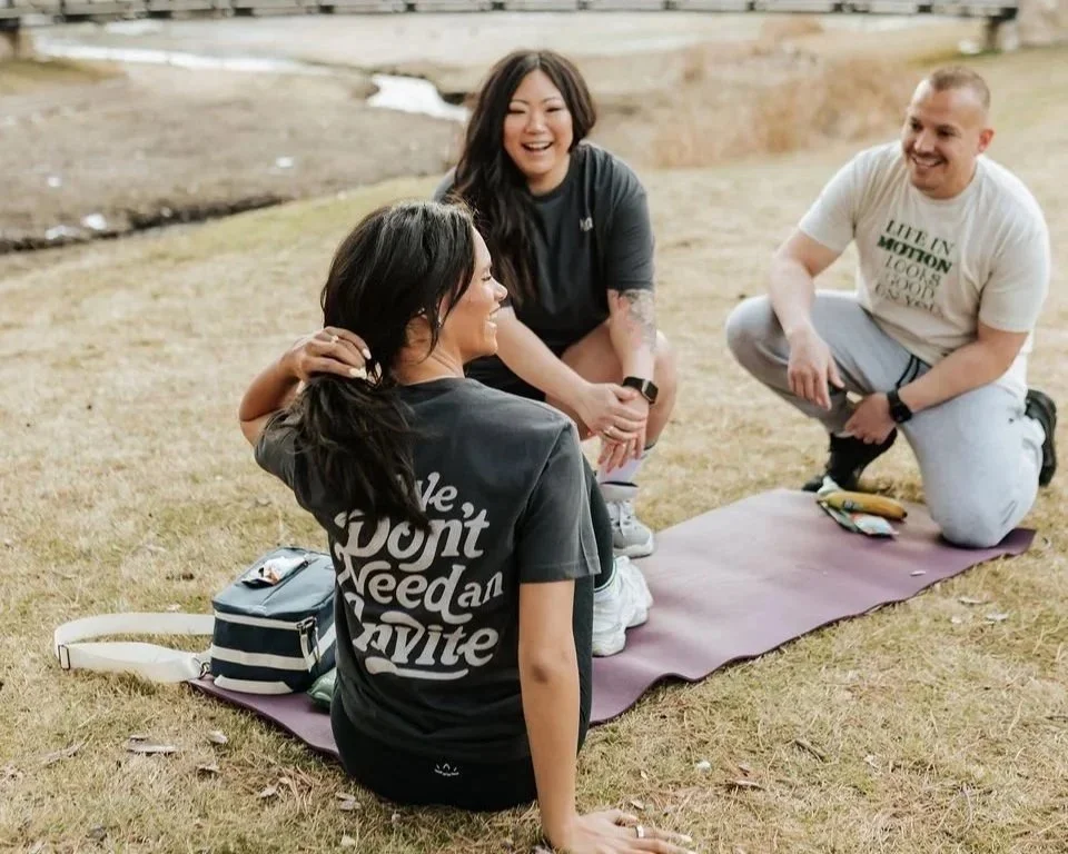 Three people sitting on the grass wearing Kanvas Movement t-shirts