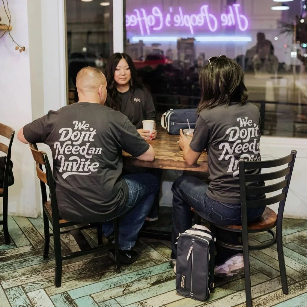 Three people sitting at a wooden table in a restaurant or cafe, all wearing matching black t-shirts with the slogan 'We Don't Need an Invite'; two are women and one is a man, with drinks in front of them. The background shows a large window with neon sign reflections.