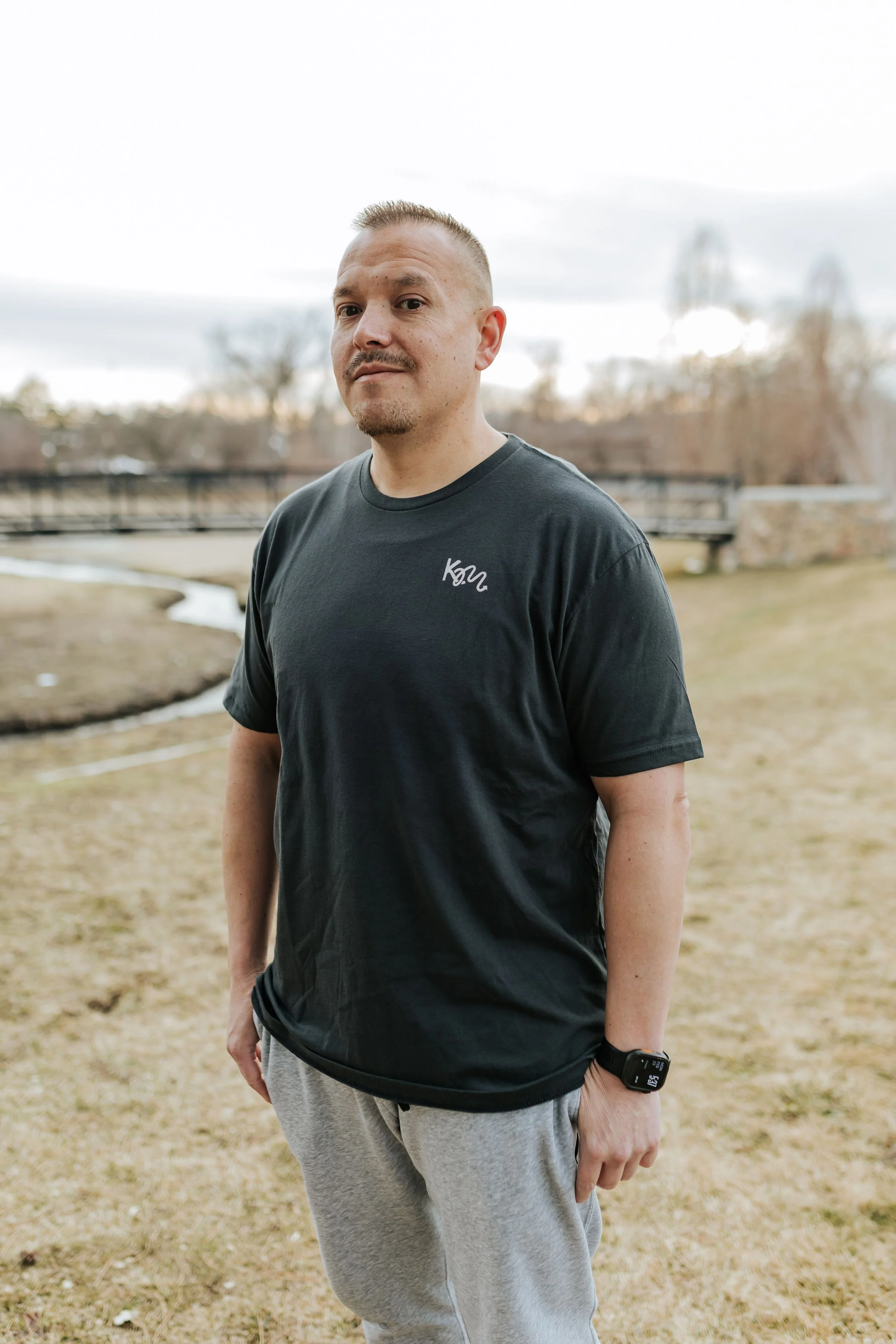 A man with short blond hair and a beard standing outdoors in a park with a bridge in the background, wearing a black t-shirt and gray sweatpants, with a smartwatch on his left wrist.