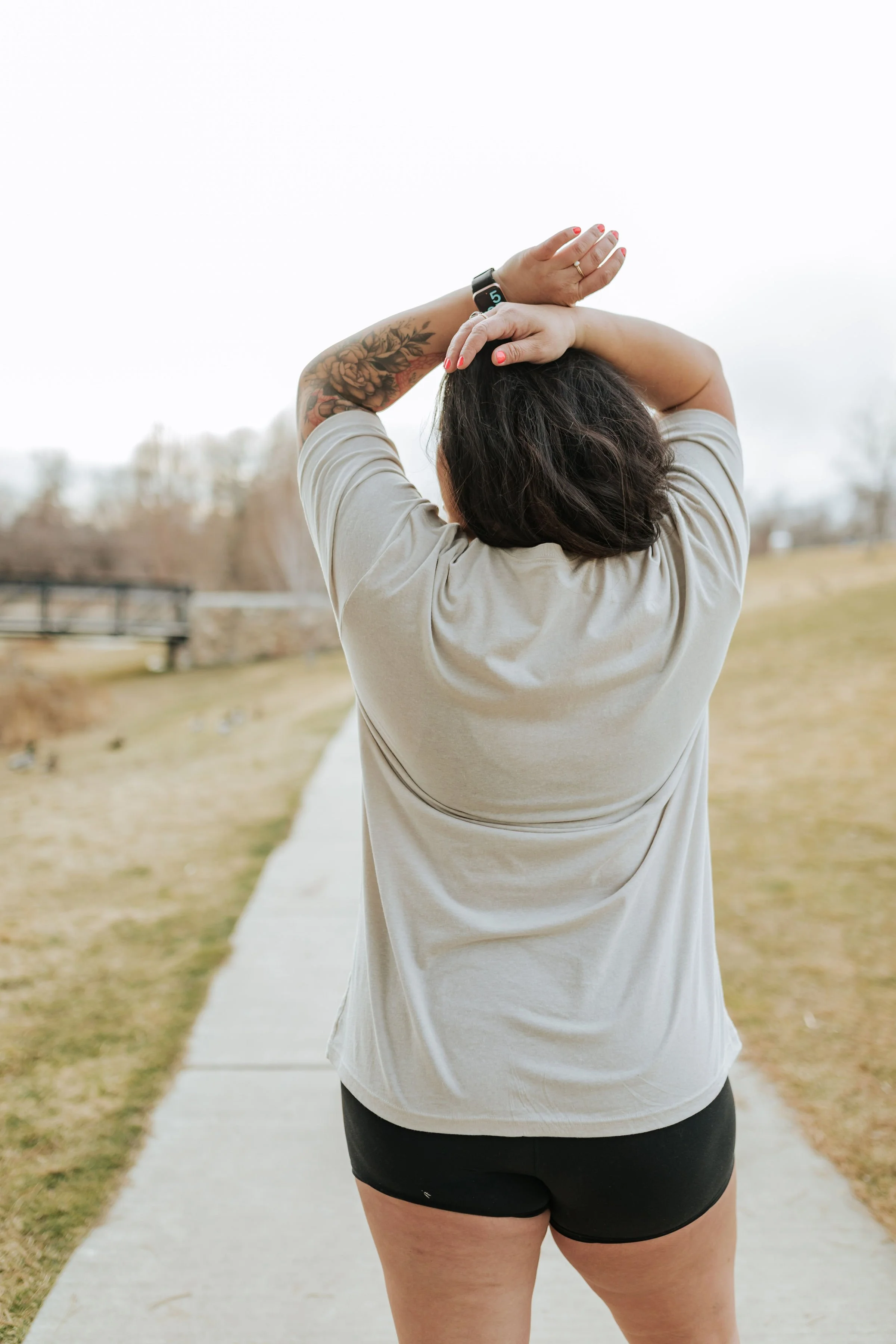 Person standing on a sidewalk outdoors stretching or relaxing with arms behind head during daytime.