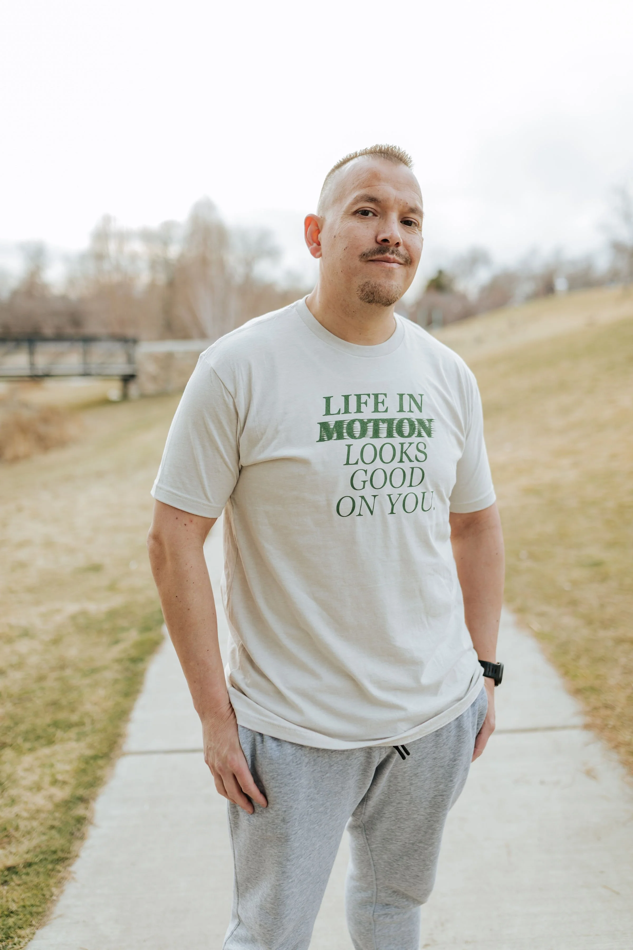 A man in casual clothing standing outdoors on a sidewalk, with trees and a bridge in the background.