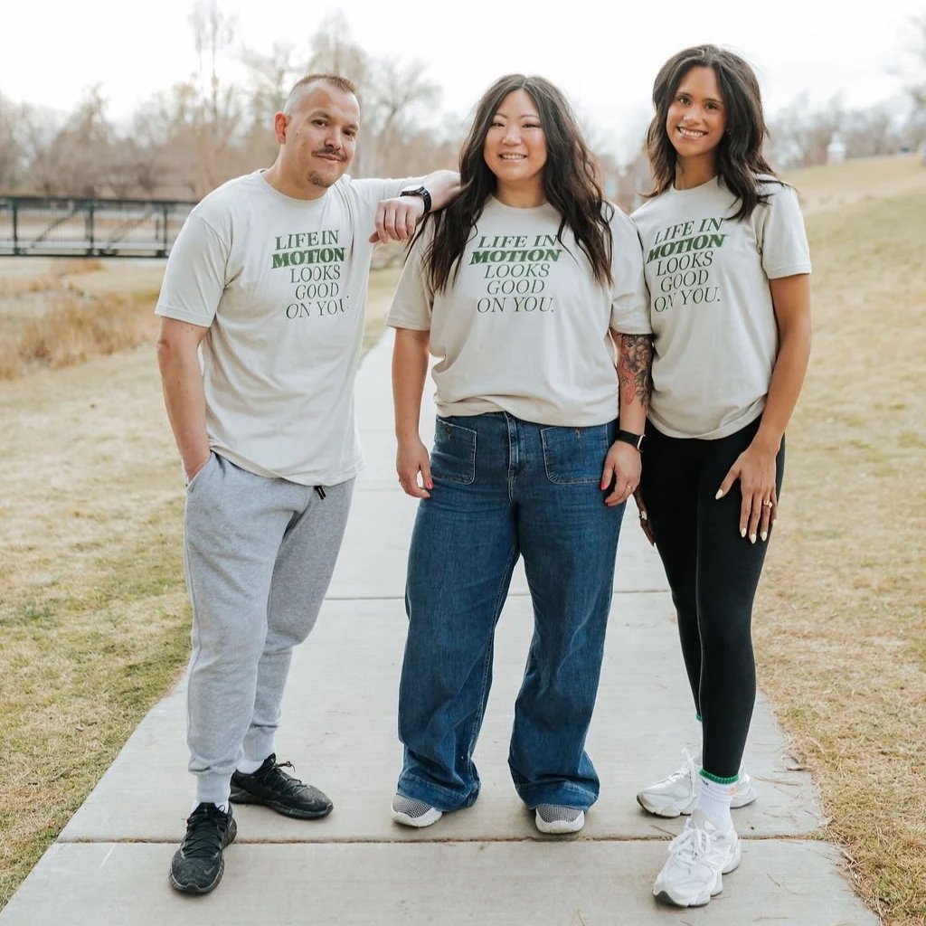 Three friends standing outdoors on a pathway, wearing matching t-shirts that say "Life in Motion Looks Good on You." They are smiling and looking at the camera, with a park-like setting in the background.