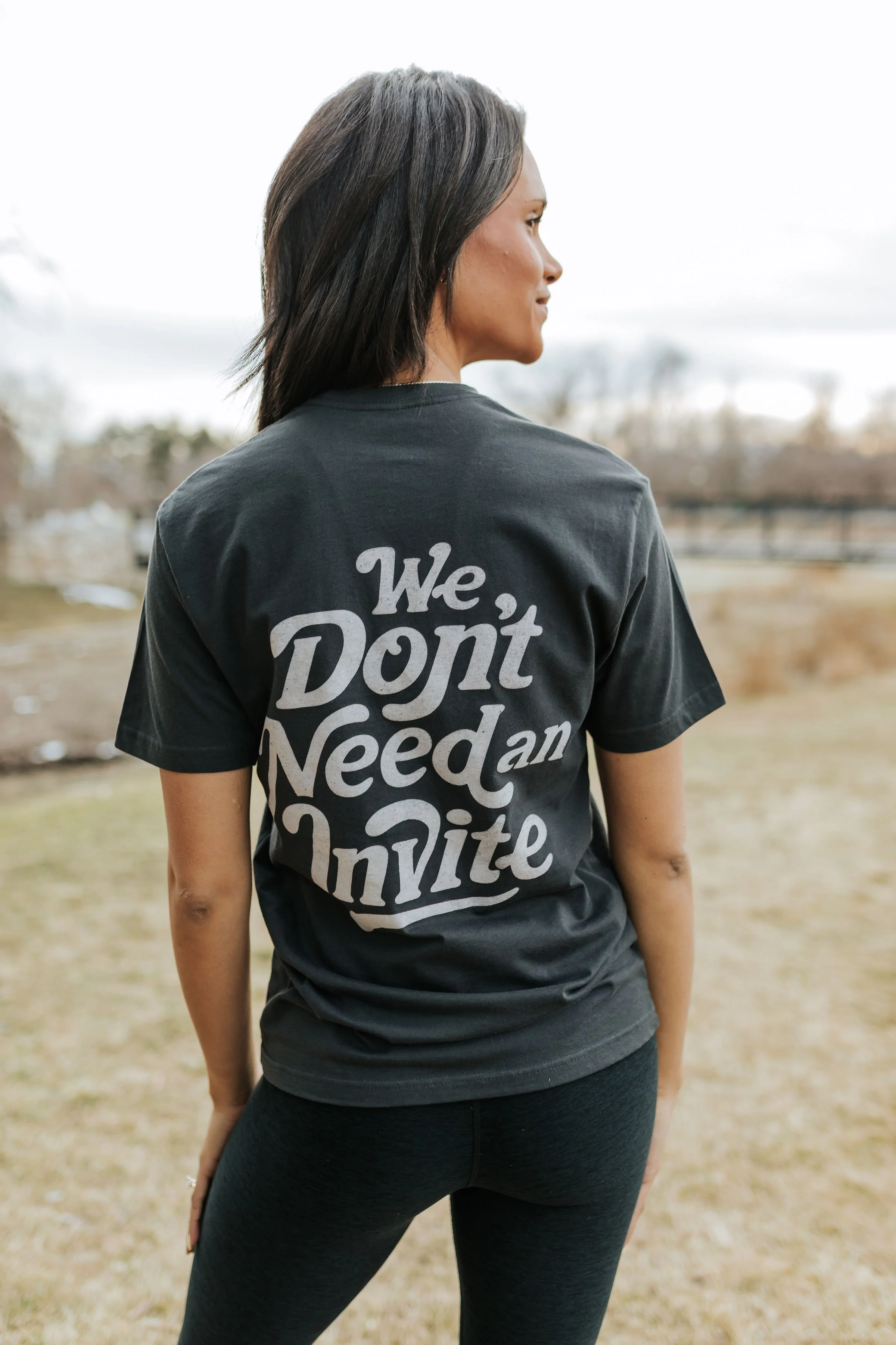 A woman standing outdoors wearing a black T-shirt with the phrase 'We Don't Need an Invite' written on the back in large white letters.