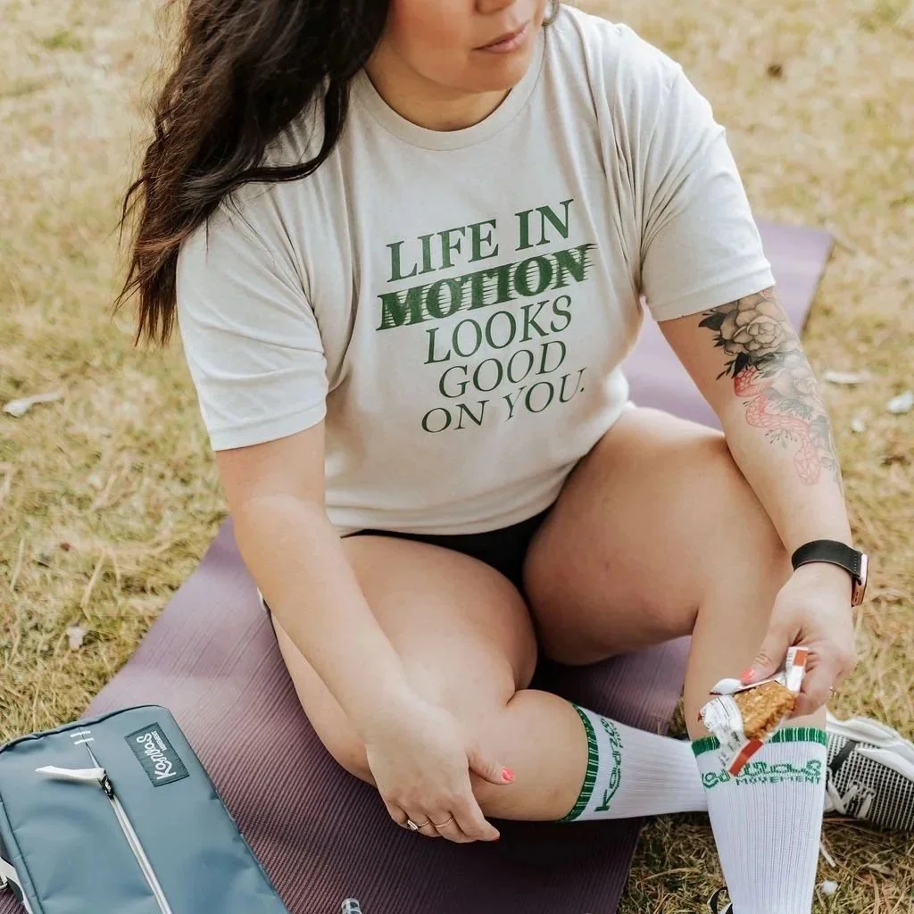 A young woman with dark hair sitting on a purple mat outdoors on grass, wearing a gray t-shirt with green text, white athletic socks, and holding a snack bar in her hand.