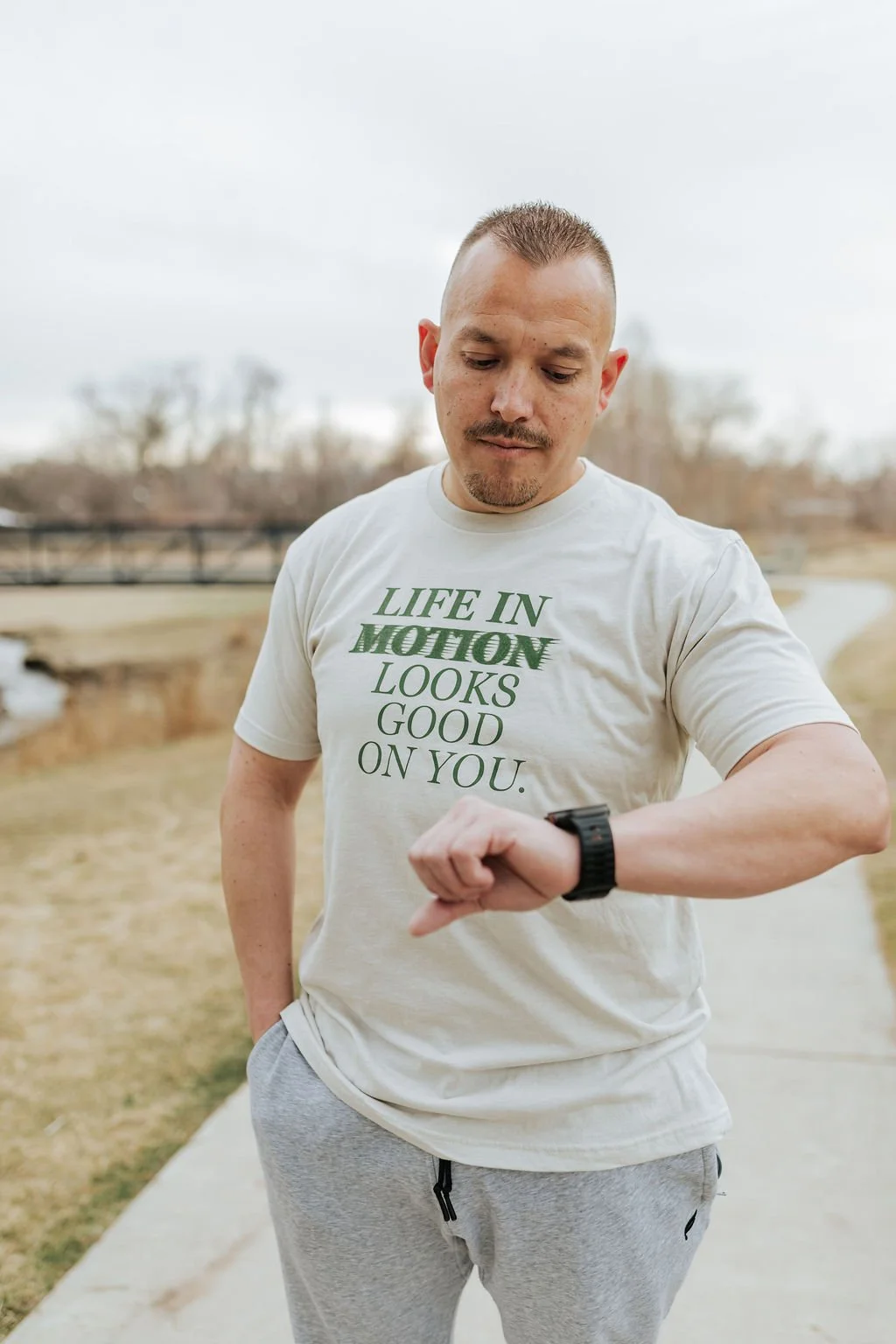 A man looking at his smartwatch outdoors on a cloudy day, wearing a white T-shirt with "Life in Motion looks good on you" text and light gray sweatpants.