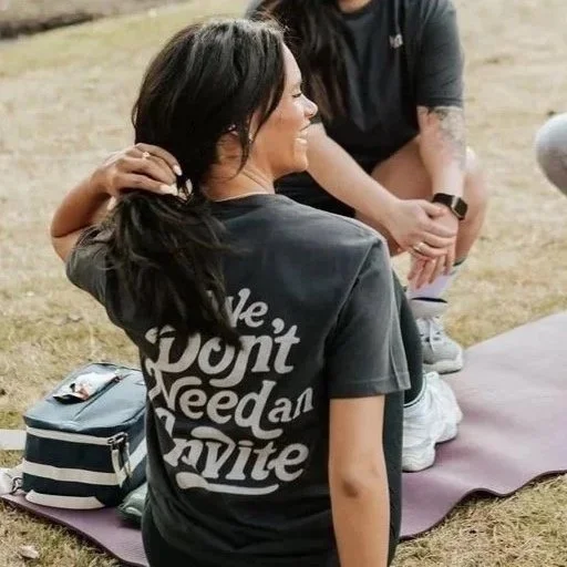 A woman with long dark hair tied in a ponytail, wearing a black T-shirt with the text 'We Don't Need an Invite', sitting on a purple yoga mat outdoors, talking to a person kneeling in front of her.