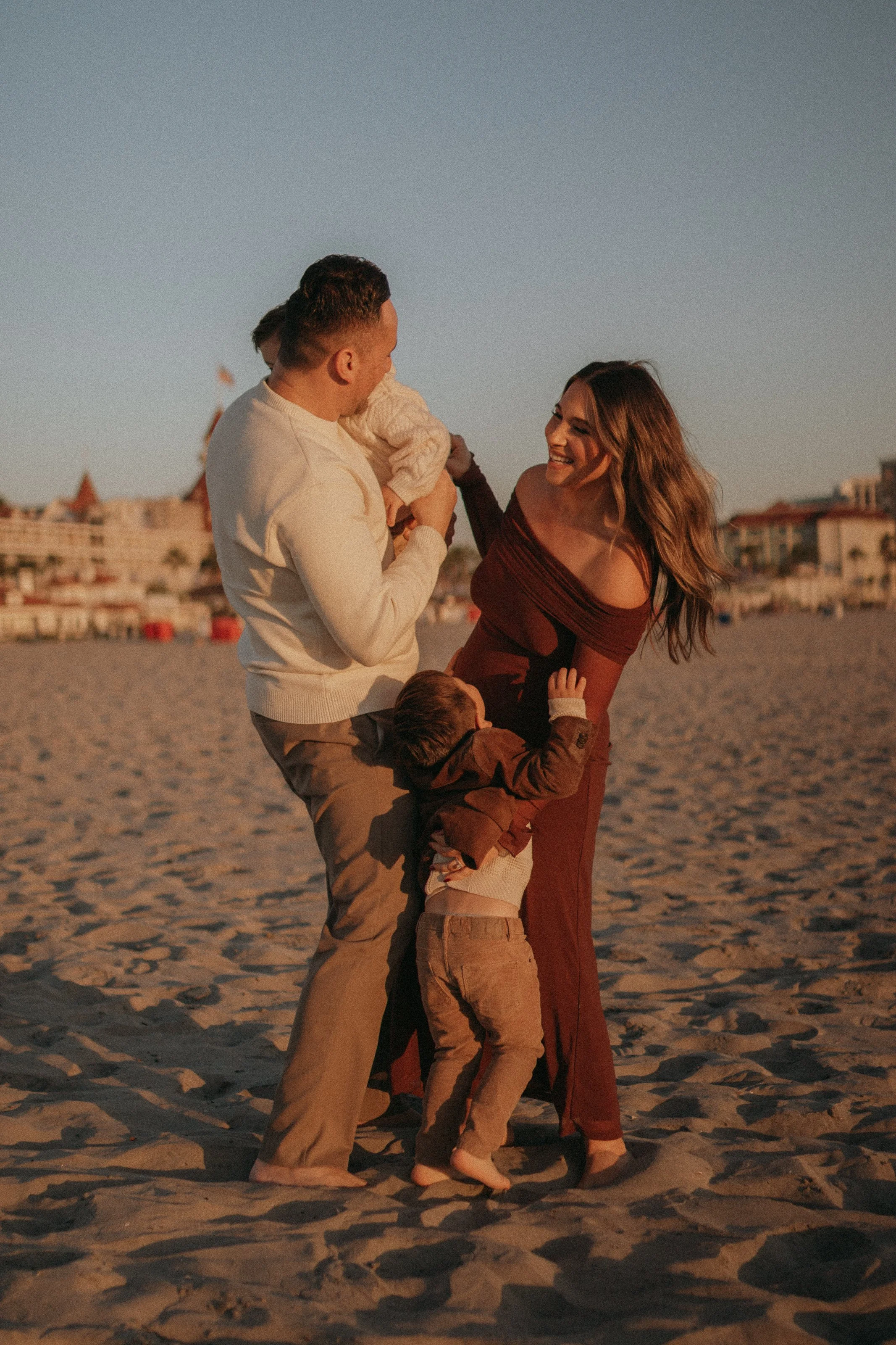 A family of four on the beach during sunset, with a father holding a baby, a smiling mother, and a young boy hugging her leg.