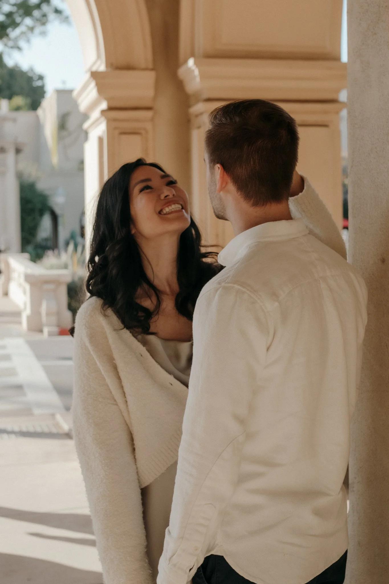 A woman and a man are smiling and looking at each other, standing close to each other outside near a building with beige columns.