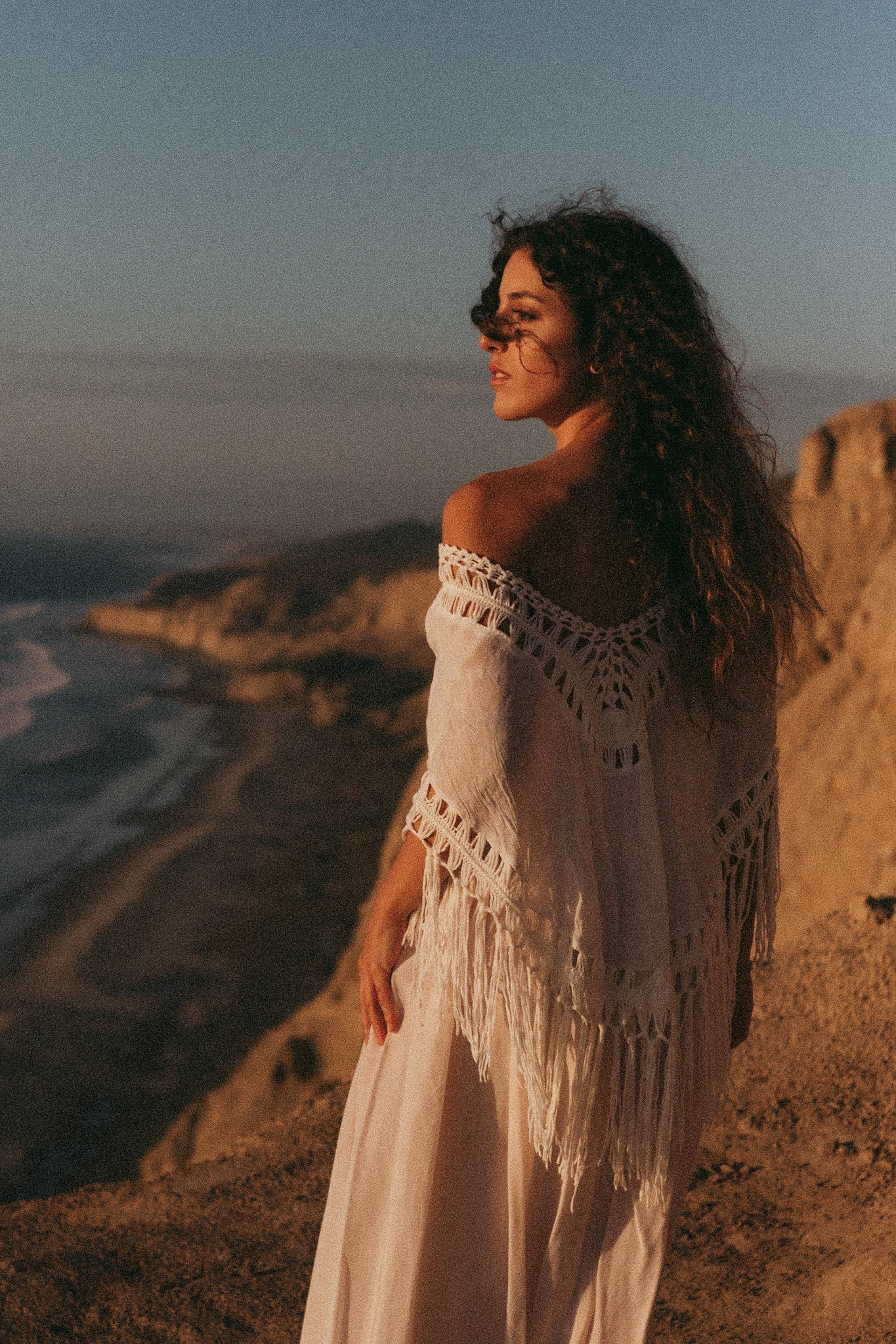 A woman with curly hair wearing a white off-shoulder dress and a fringe shawl standing on a sandy cliffside overlooking the ocean during sunset.
