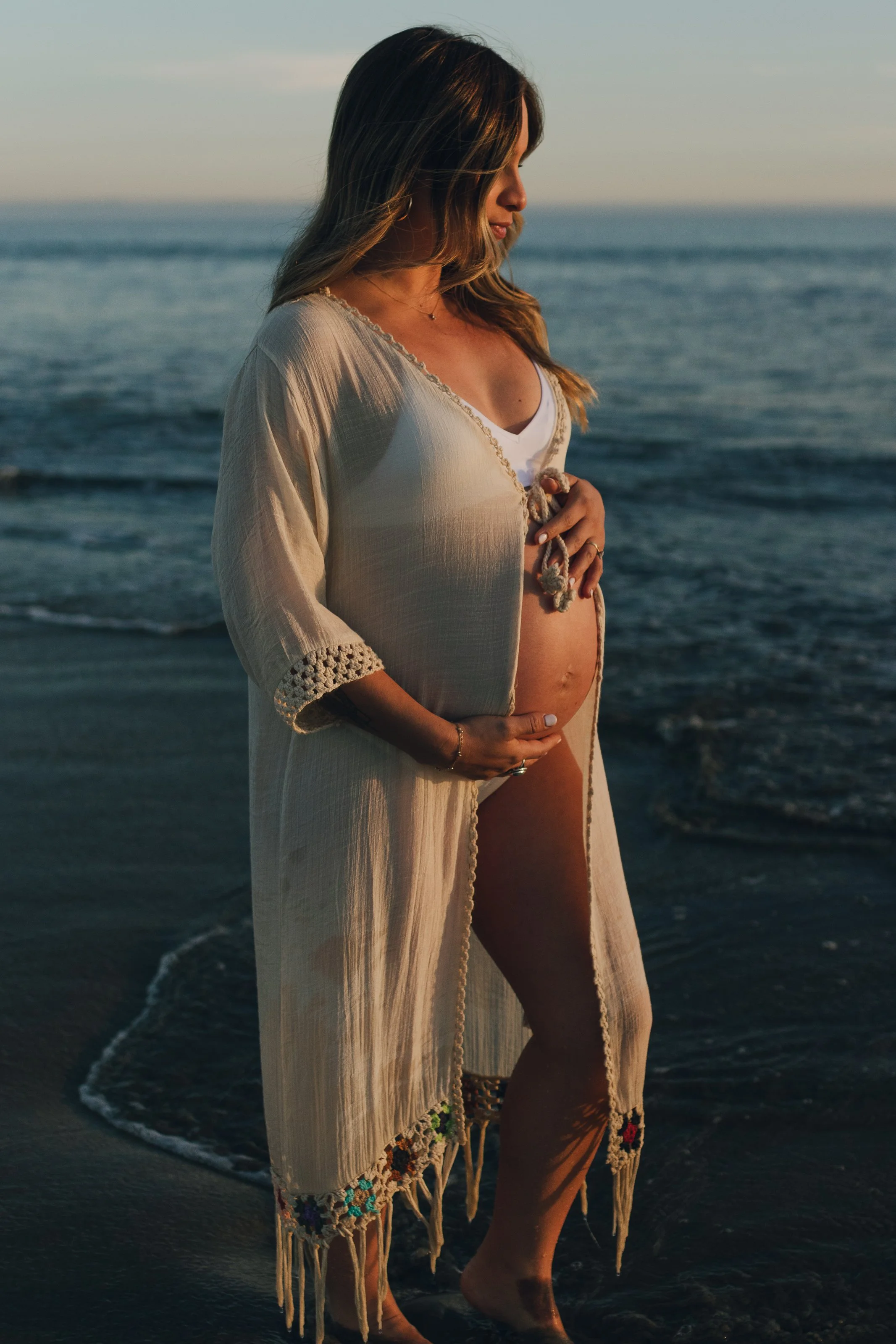 A pregnant woman standing on a beach at sunset, gently holding her belly with one hand and touching her shirt with the other, wearing a light, embroidered cover-up and a white top underneath.