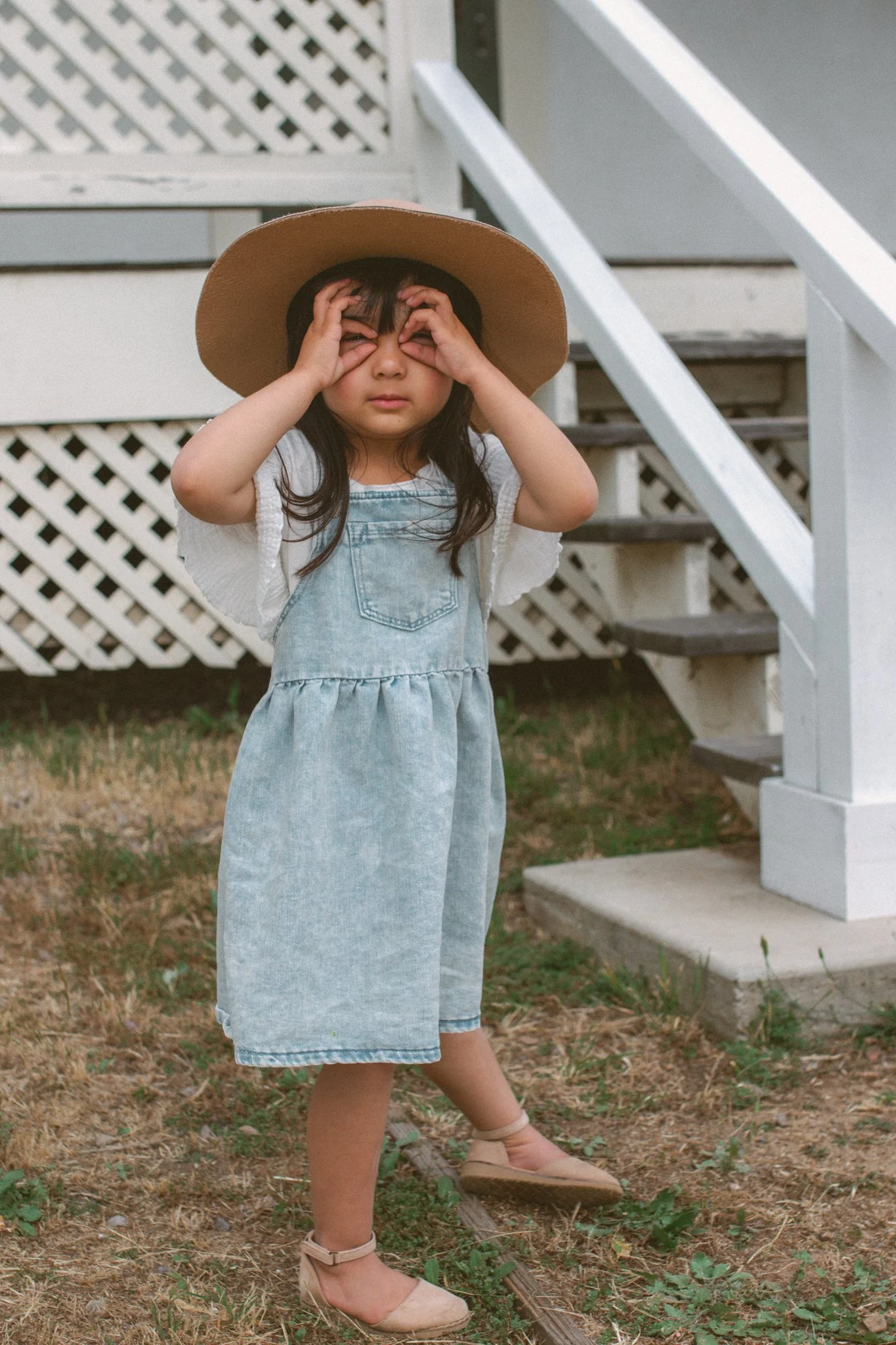 Young girl wearing a large sun hat and denim dress, standing outdoors near a white house staircase, holding her face with her hands.