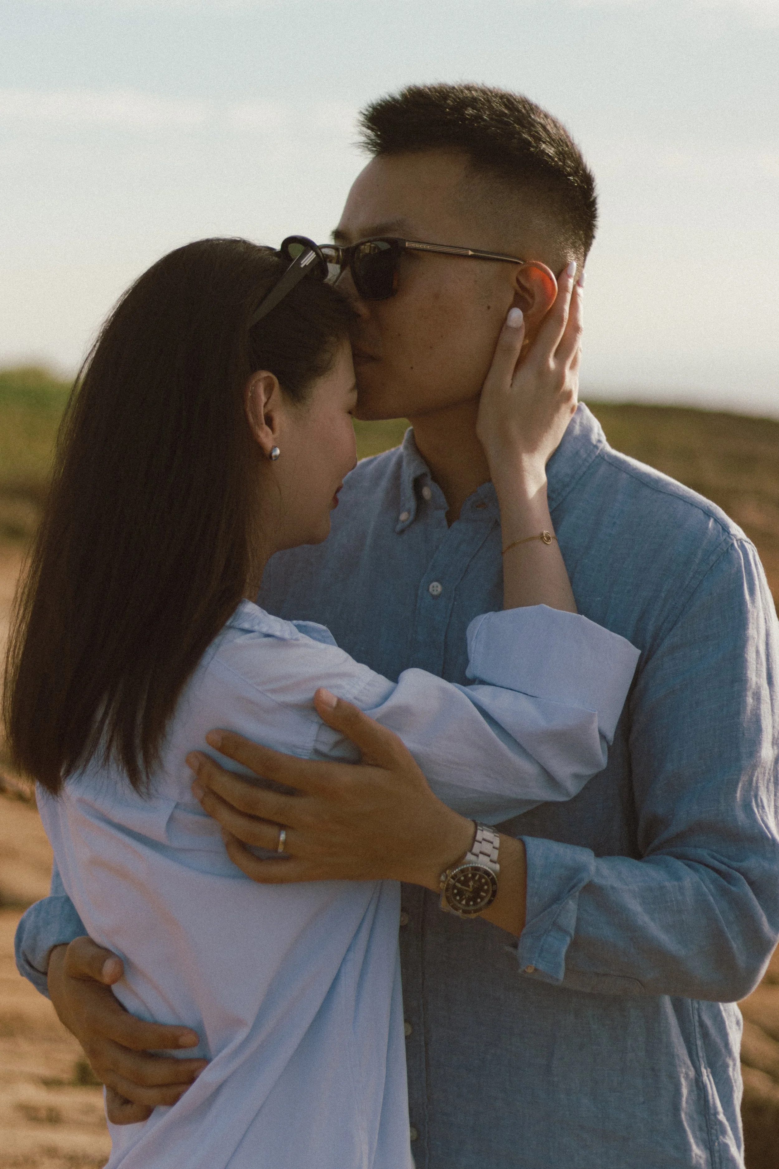 A couple embracing outdoors, with the man kissing the woman's forehead.