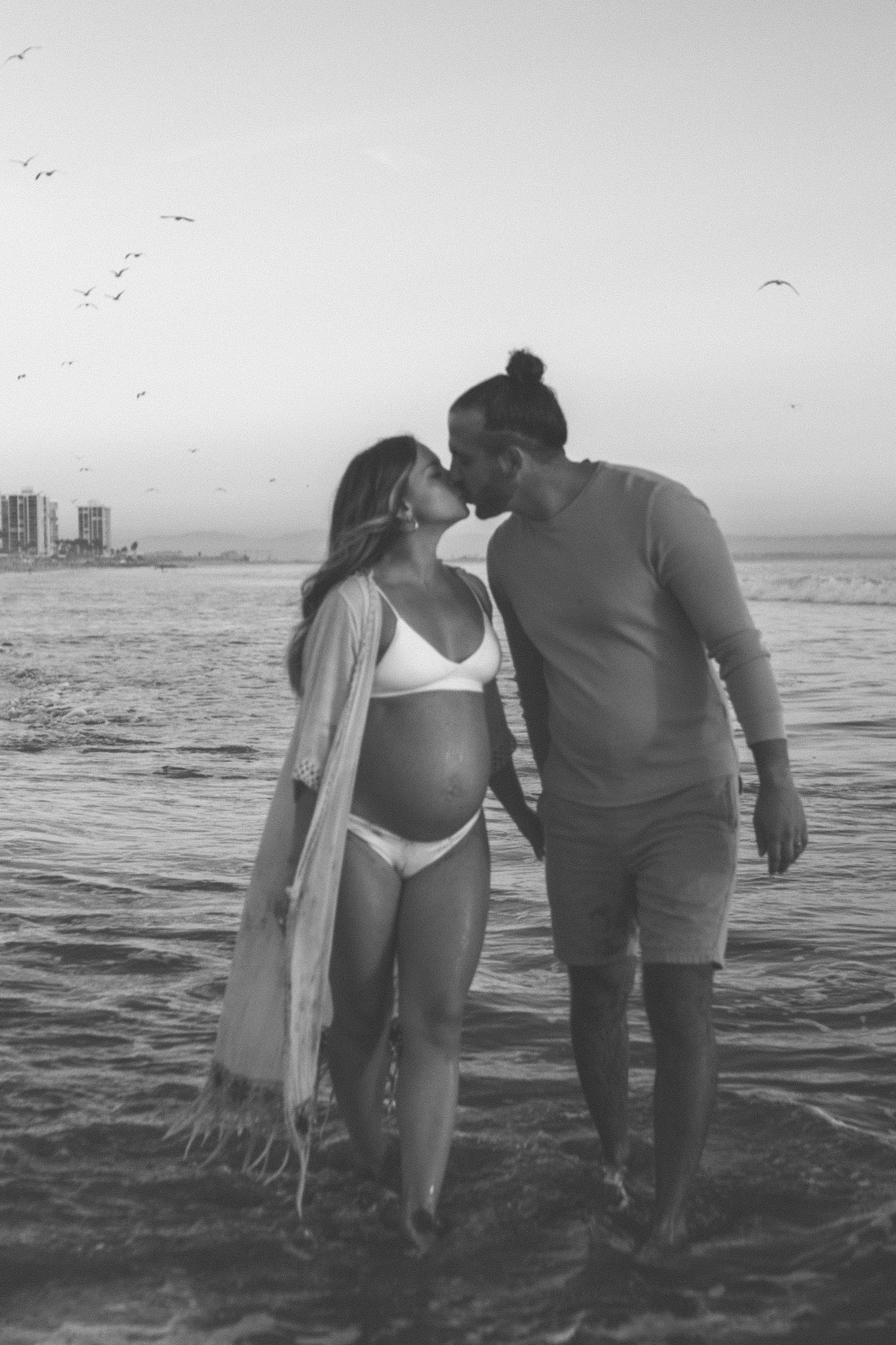A pregnant woman and a man sharing a kiss on the beach at sunset, with the ocean and city buildings in the background.