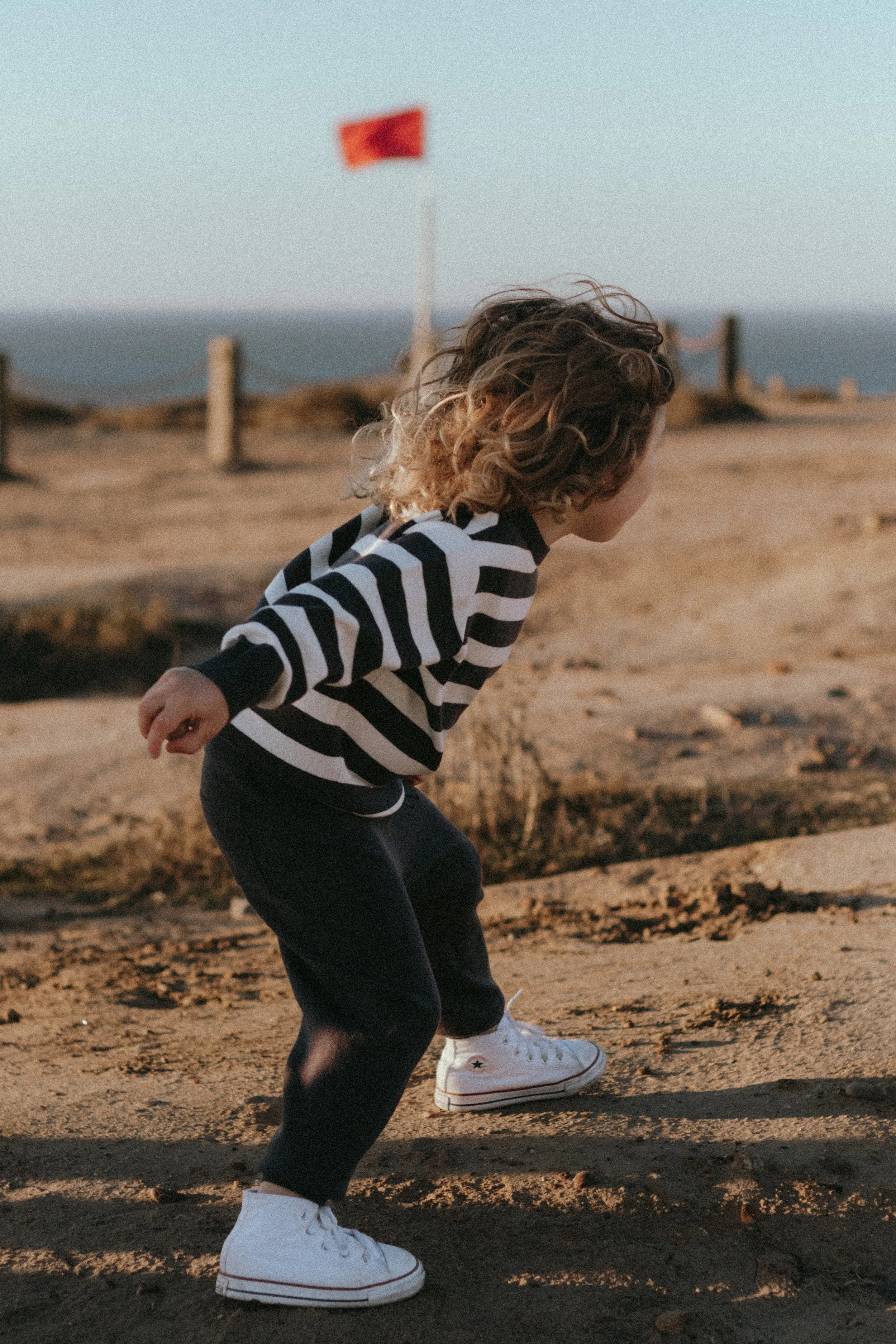 Child with curly hair wearing striped shirt and white sneakers, playing outdoors on a sandy path near the coast with a red flag in the background.