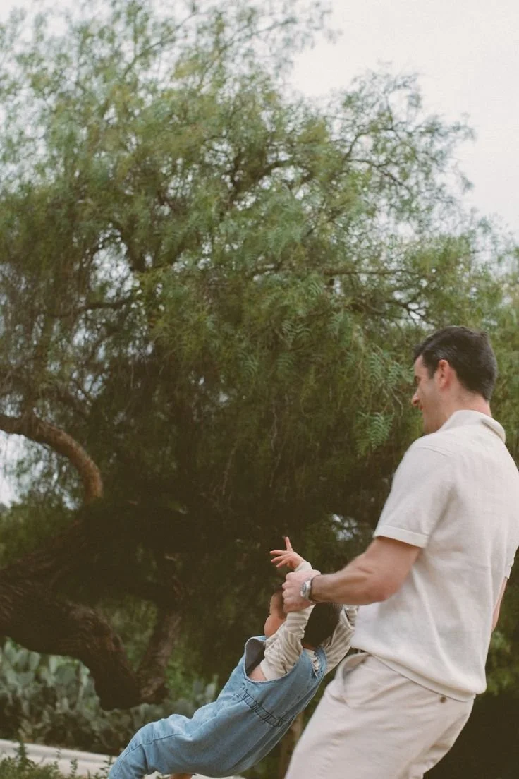 A man and a young girl playing outdoors near a large tree, with the girl hanging upside down by her legs and the man holding her hands.